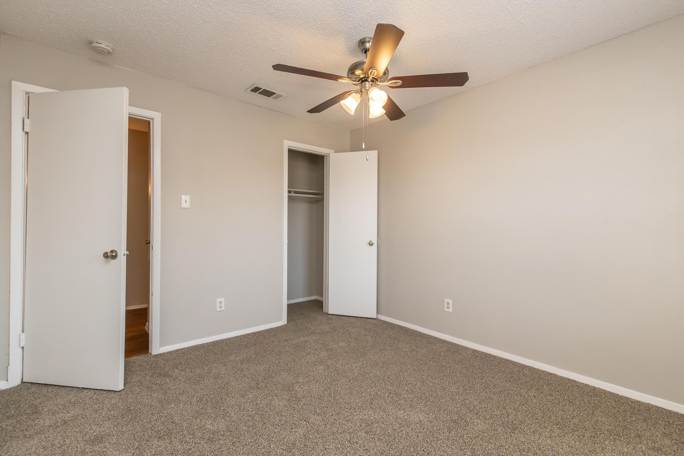 A small, empty room with light gray walls and carpeted flooring. There is a ceiling fan with five blades in the center. One door leads to a closet, while another door is slightly ajar, showing a glimpse of a hallway. Natural light enters through the open door, creating a bright atmosphere.