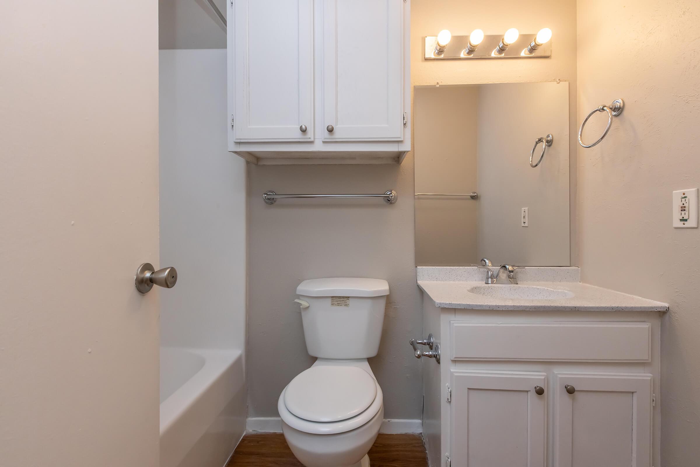A small bathroom featuring a white toilet, a sink with a granite countertop, and a large mirror above it. There's a shower/tub combination and a linen cabinet above the toilet. The walls are painted light beige, and the space is well-lit with overhead lights.