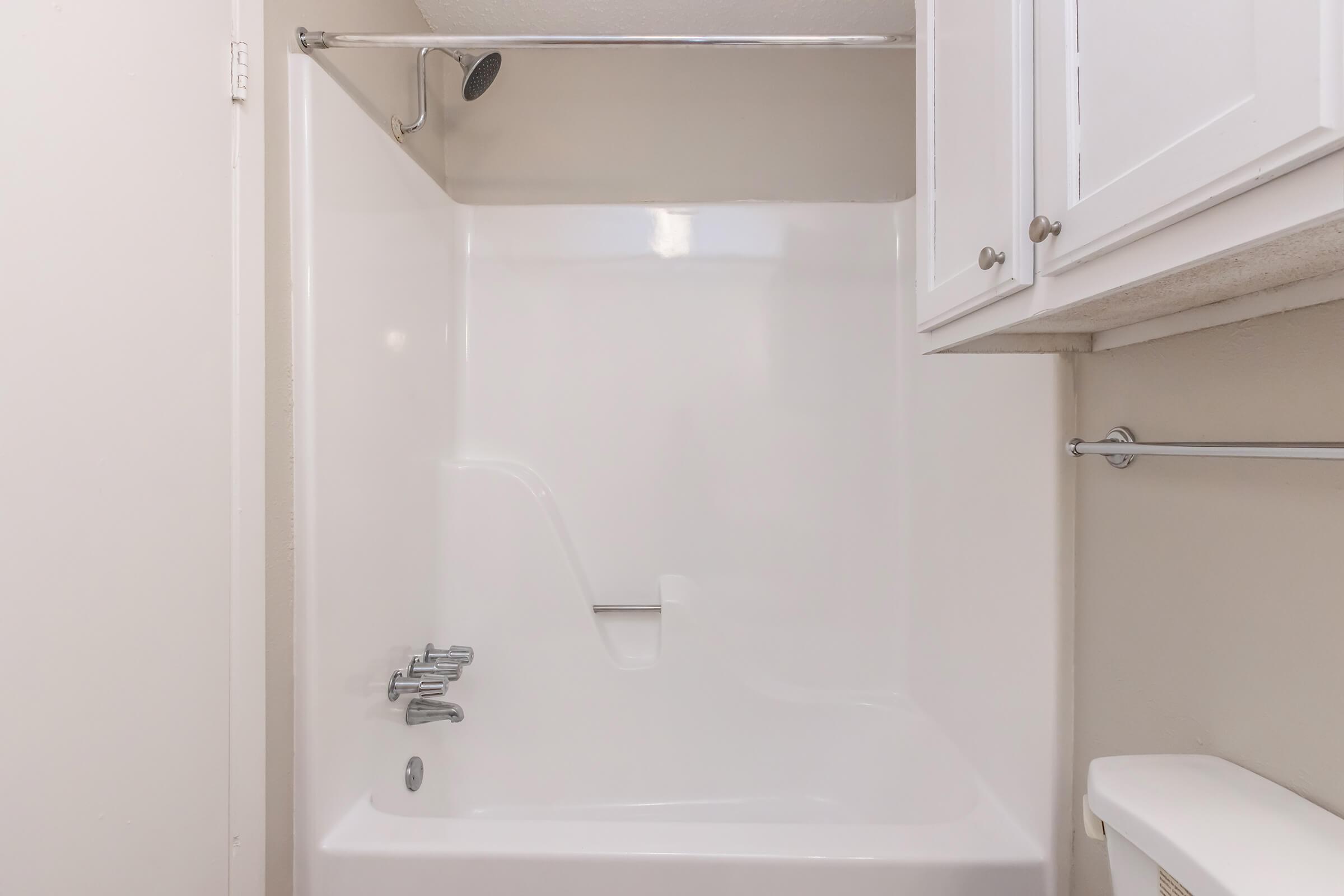 A clean, well-lit bathroom featuring a white fiberglass tub with a showerhead. The wall is painted a light color, and there are white cabinets above. A towel rack is mounted on the opposite wall, creating a simple and functional space.