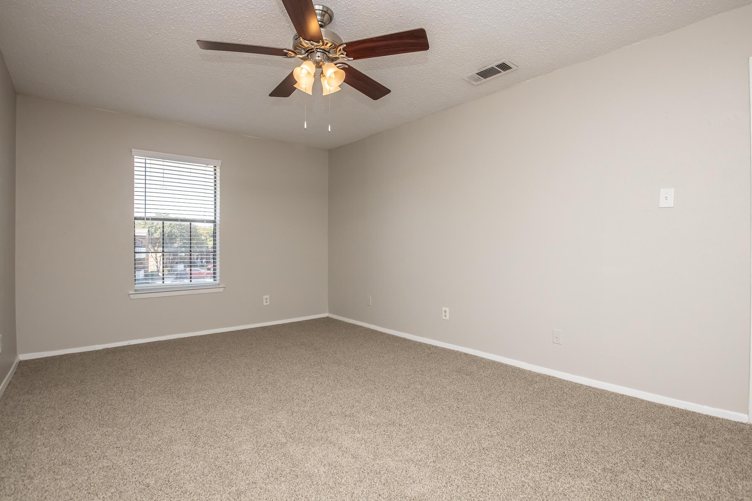 A spacious, empty room with beige carpet and light gray walls. A ceiling fan with four light fixtures hangs from the ceiling. There is a window on the left side with blinds, allowing natural light to enter. The space is well-lit and clean, providing a blank canvas for decorating.