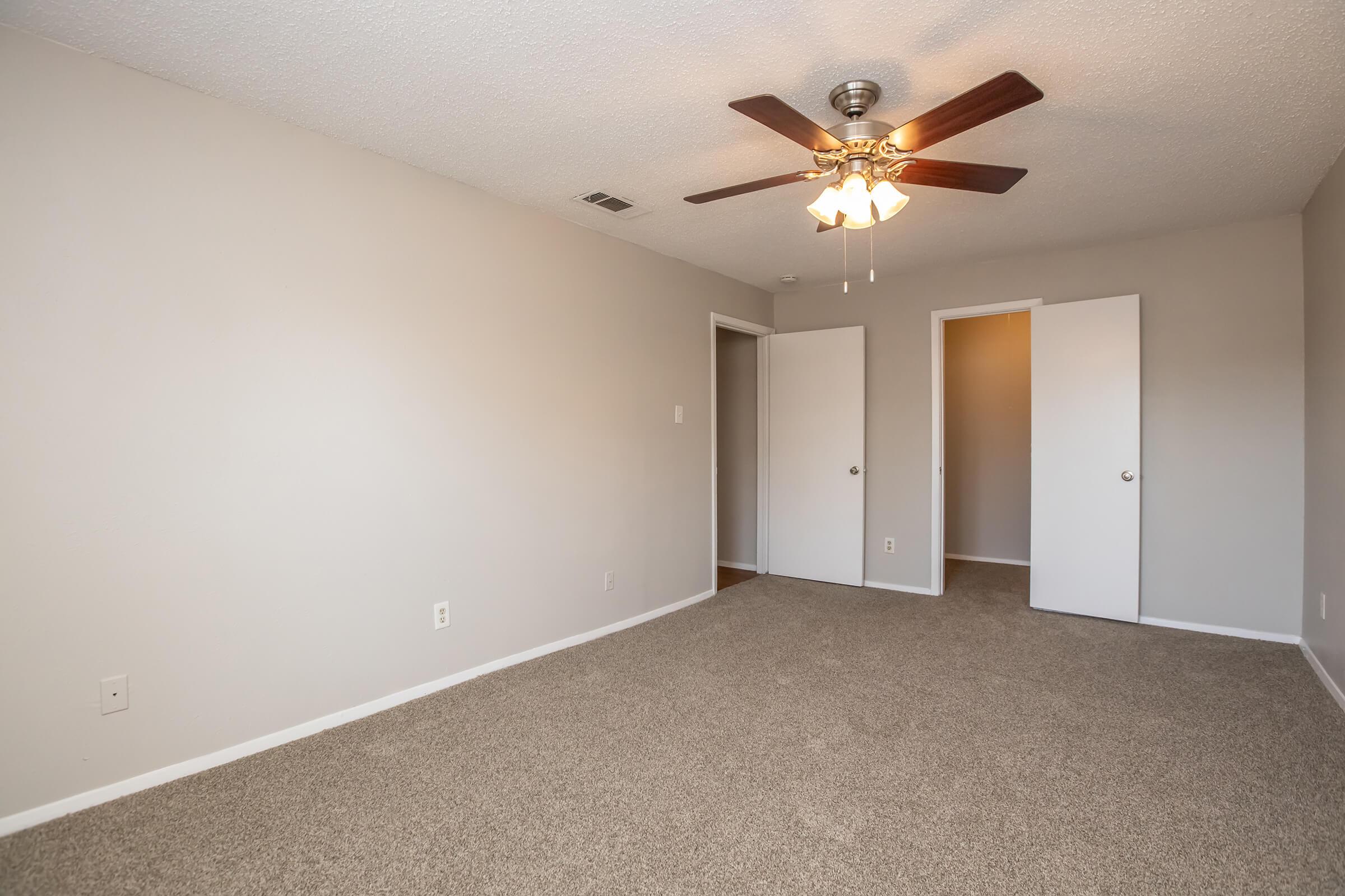 A spacious room with light gray walls and soft carpeting. There is a ceiling fan with a light fixture, and two closed closet doors in the background. The room is well-lit, featuring a neutral color palette and an empty, clean appearance, suitable for various uses.