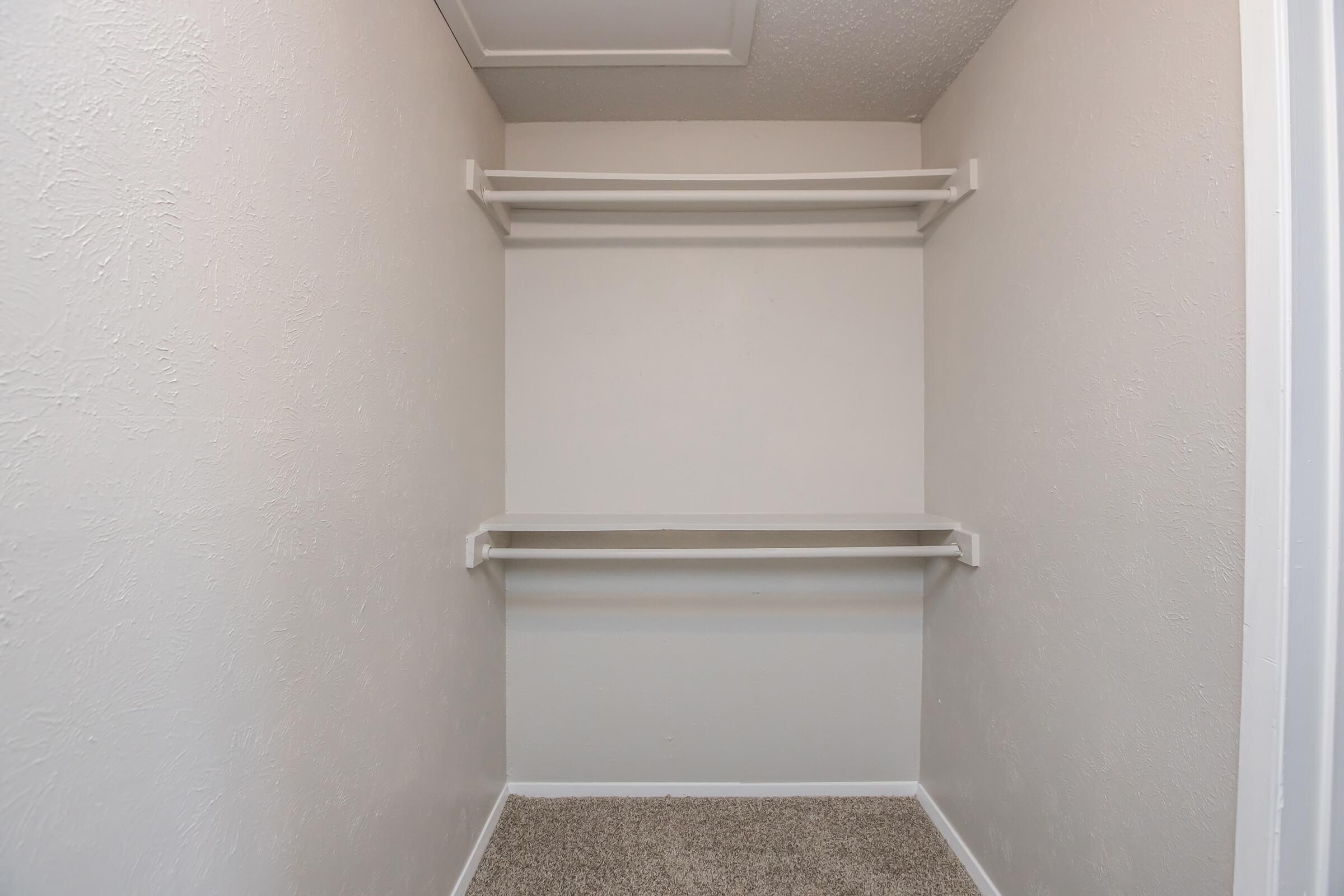 A clean, empty closet with beige walls and two white shelves. The floor is covered in light-colored carpet, and there is a ceiling panel visible above. The overall appearance is simple and uncluttered, suggesting a fresh space ready for organization.