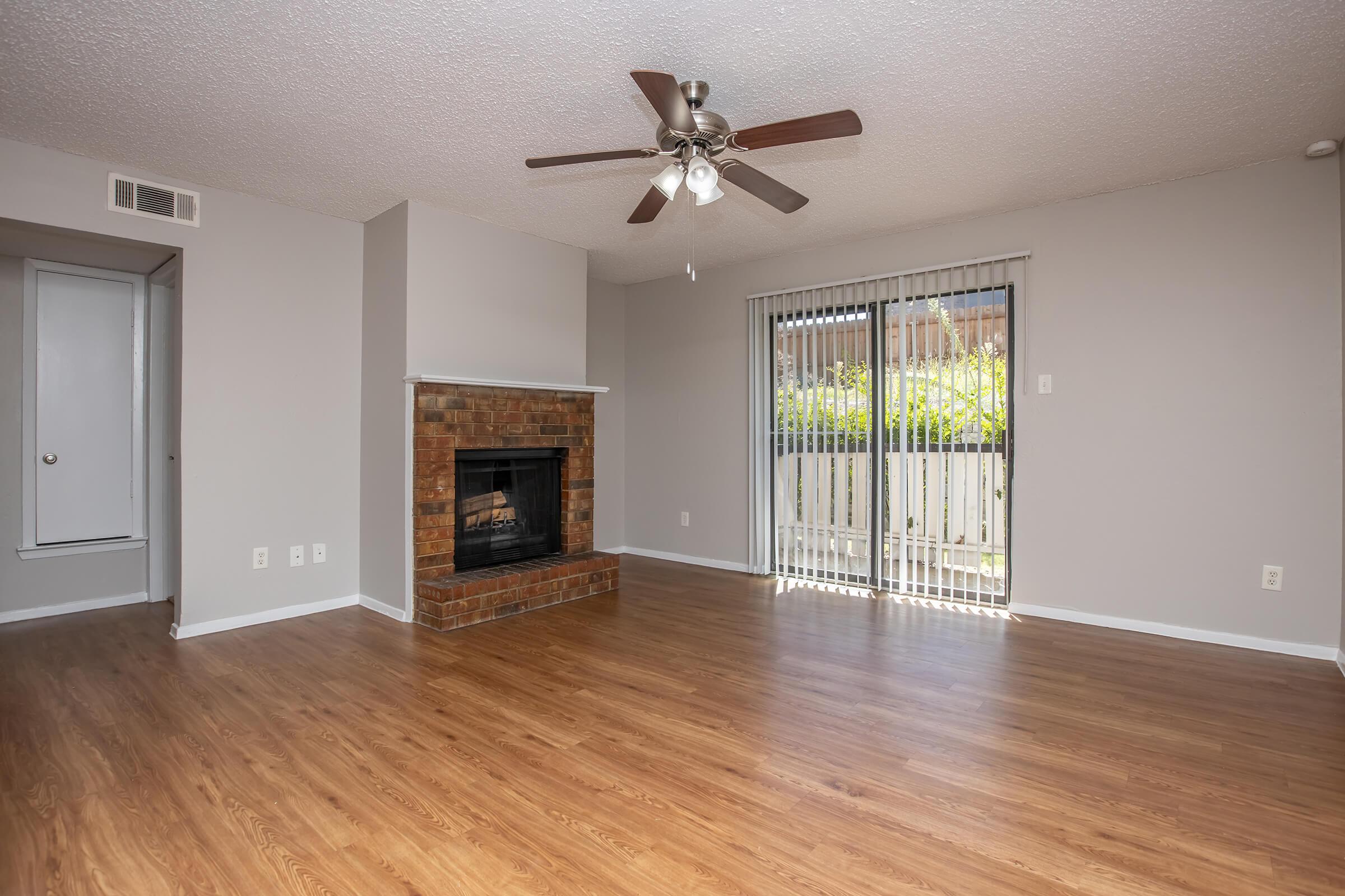 A spacious living room featuring a ceiling fan, a brick fireplace, and large sliding glass doors that open to an outdoor area. The walls are painted light gray, and the floor is covered in laminate wood. Natural light fills the space, creating a bright and inviting atmosphere.