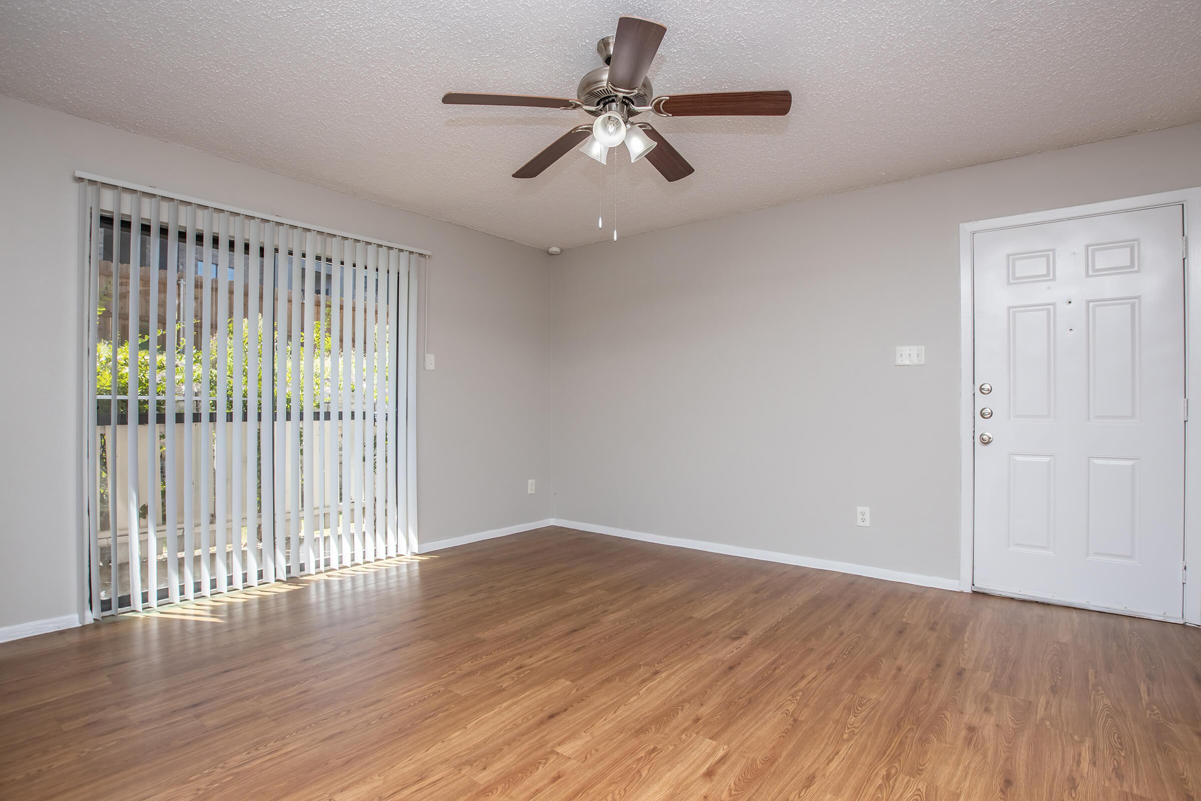 A spacious, empty room with light brown wooden flooring, a ceiling fan with light fixtures, and a large window with vertical blinds. The wall is painted a soft gray, and there is a white door on the right. Natural light streams in from the window, creating a bright and welcoming atmosphere.