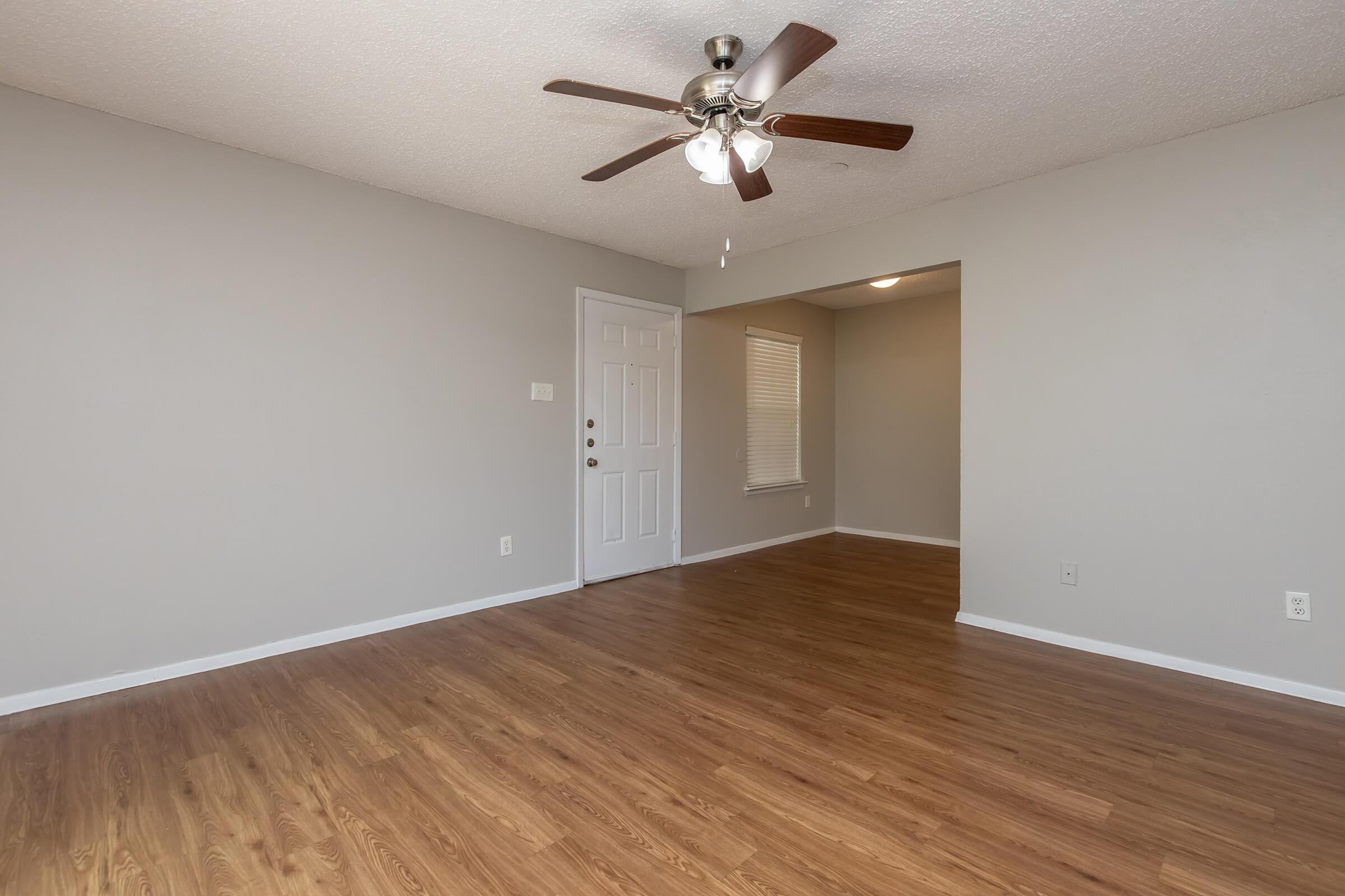 A spacious room with light brown laminate flooring, ceiling fan with lights, and gray walls. There is a white door and a window on one side, providing natural light. The layout appears open, leading into another area.