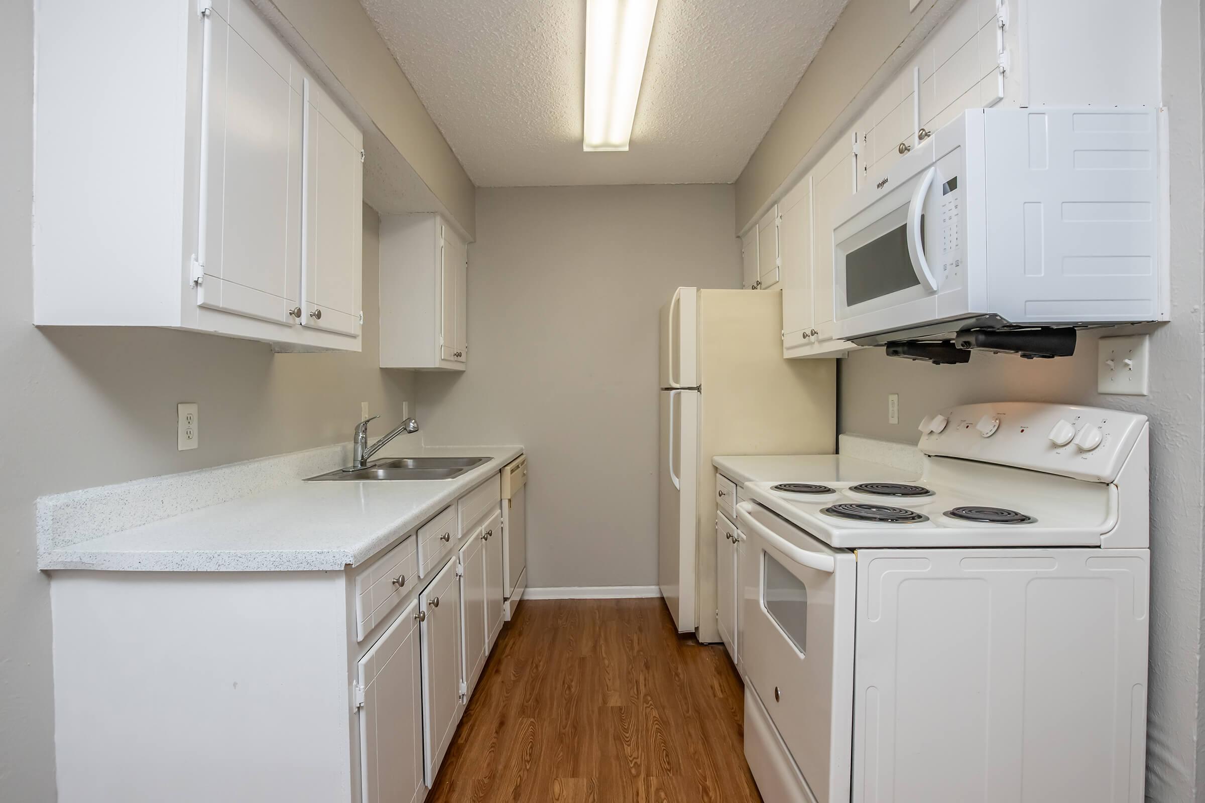 A modern kitchen featuring white cabinetry, a stainless steel sink, a microwave above the stove, and a refrigerator. The countertops are light-colored, and the floor is laminate wood. Soft overhead lighting illuminates the space, creating a clean and inviting atmosphere.