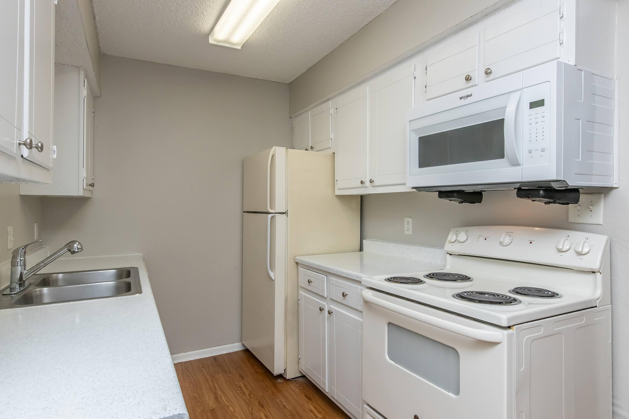 A modern kitchen featuring white cabinetry, a refrigerator, a microwave above the stove, and an electric range with a smooth countertop. The walls are painted a neutral color, and there is a single sink with a chrome faucet. The flooring is light wood laminate.