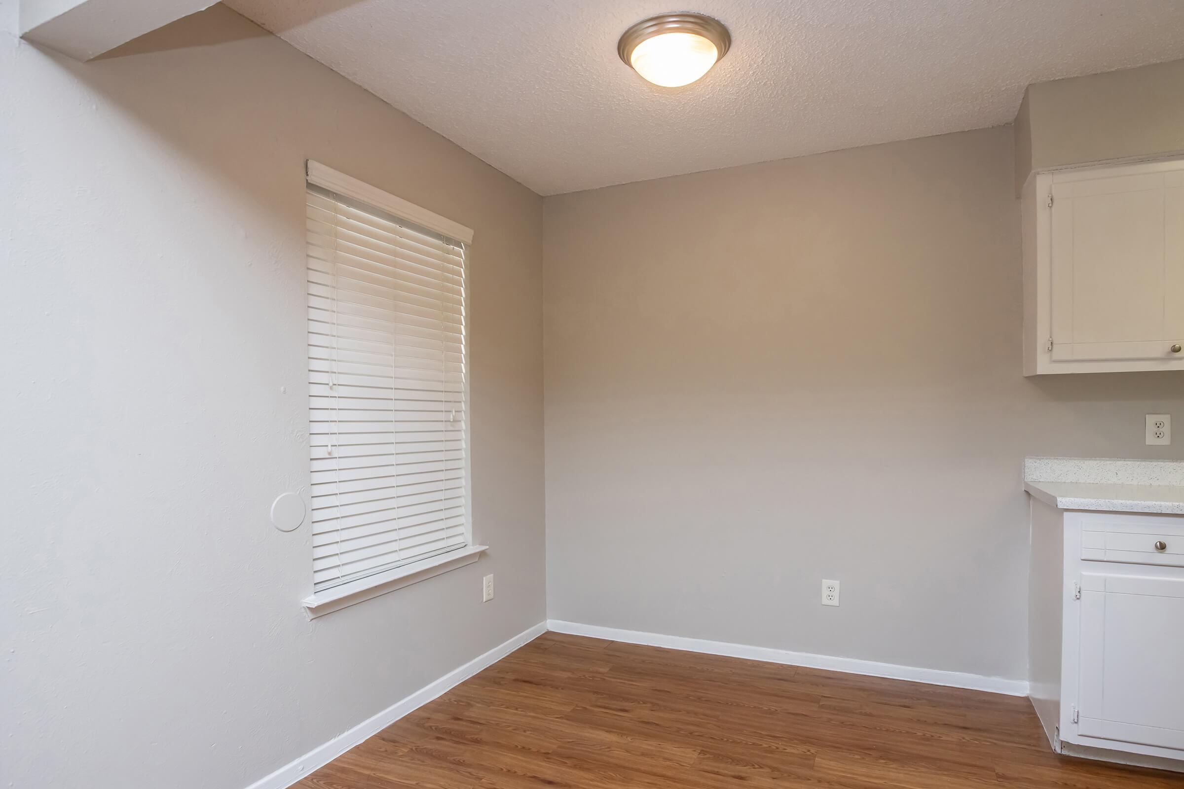 A well-lit, empty kitchen corner with light gray walls and hardwood flooring. A white window with blinds is on the left, and a small white cabinet is visible on the right against the wall. The ceiling features a simple light fixture, creating a modern and clean aesthetic.