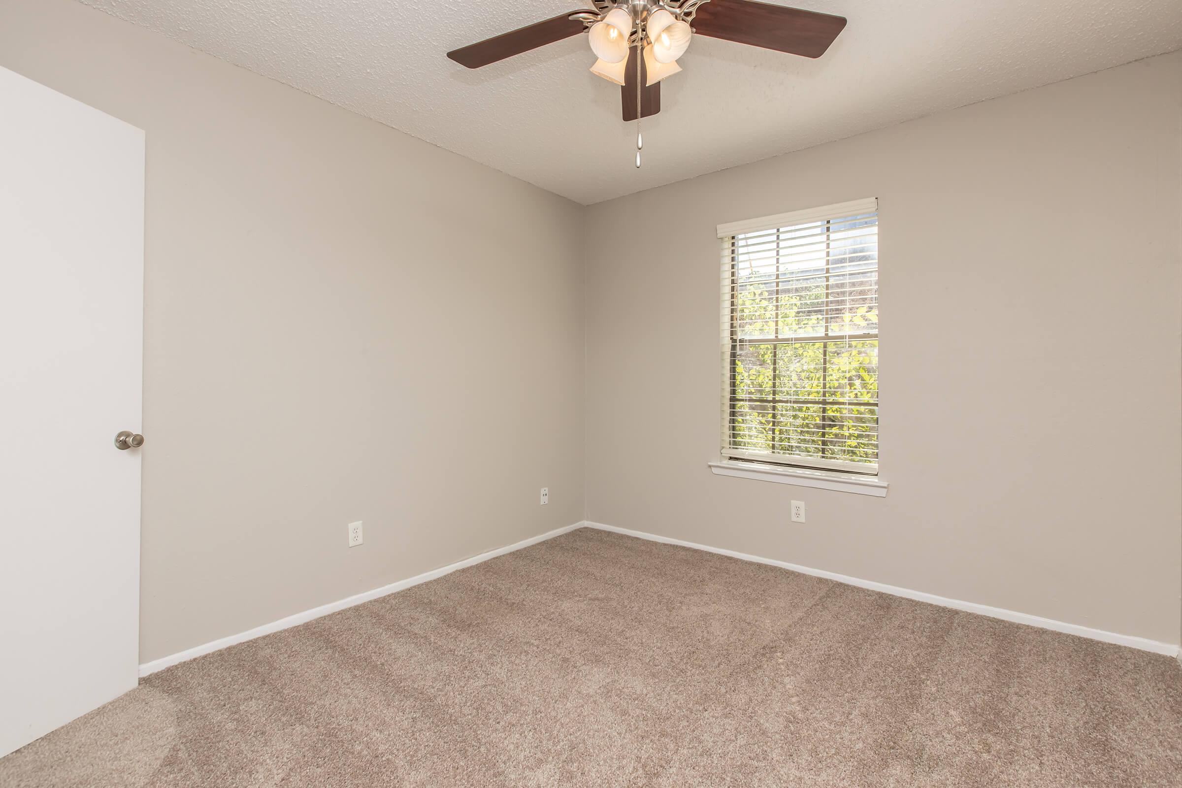 A small, empty room with light gray walls and beige carpet. It features a ceiling fan with light fixtures, a single window with blinds allowing natural light, and a white door on the left. The room appears clean and spacious, ideal for various uses or as a bedroom.