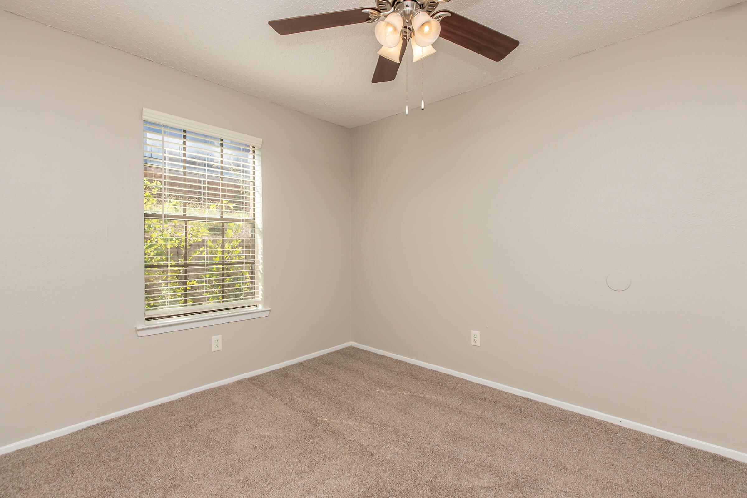 A vacant room with light beige walls and a carpeted floor. There is a window with white blinds letting in natural light, and a ceiling fan with three lights is fixed to the ceiling. The room appears empty and ready for furnishing, with minimal decoration.