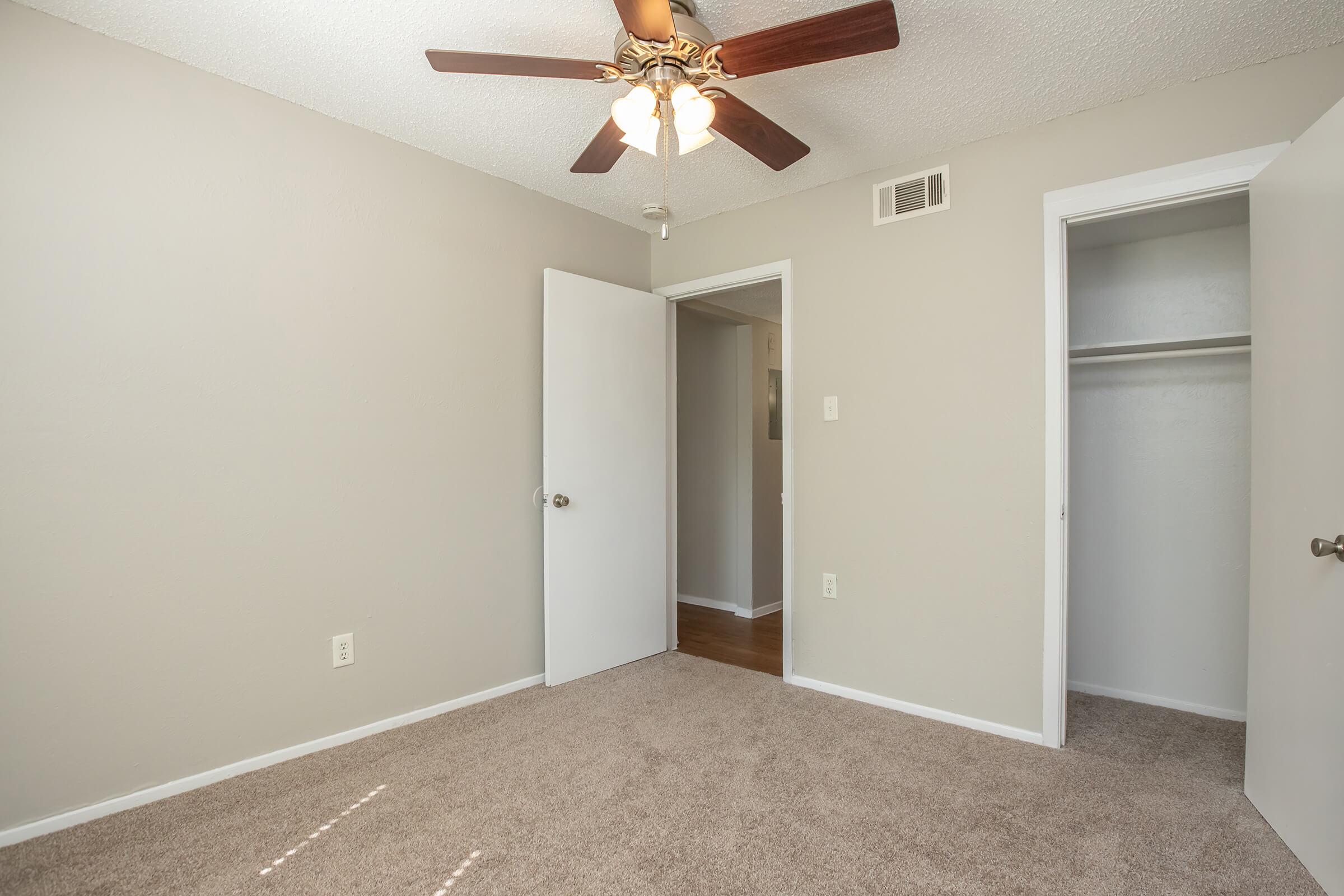 A small, empty room with beige walls and plush carpet. It features a ceiling fan with wooden blades, a closed door leading to another room, and a closet door on the right. Natural light enters through a window, creating a bright and inviting atmosphere.