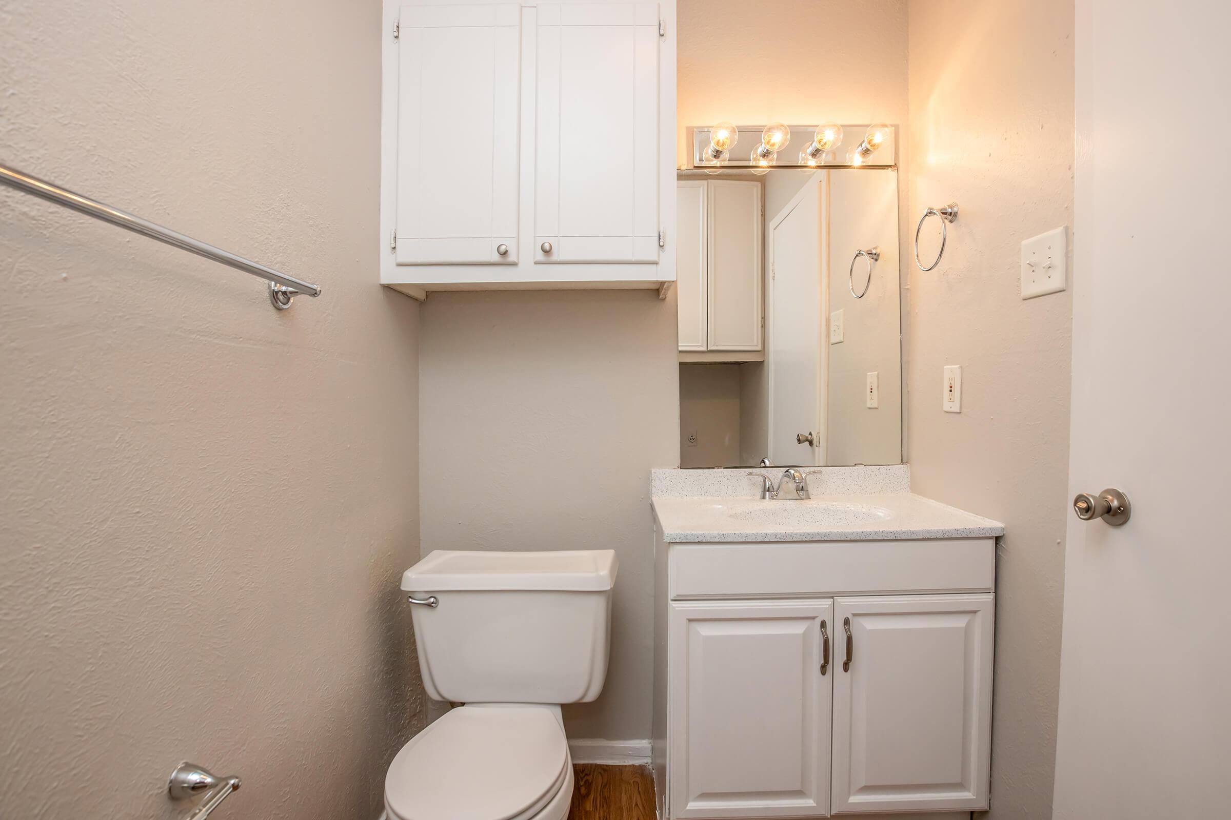 A small, modern bathroom featuring a white toilet, a white vanity with a sink, and a mirror above. The walls are painted in a light beige color, and there are light fixtures above the mirror. A cabinet is located above the vanity for storage, and the floor is wooden. Overall, the space is clean and well-lit.