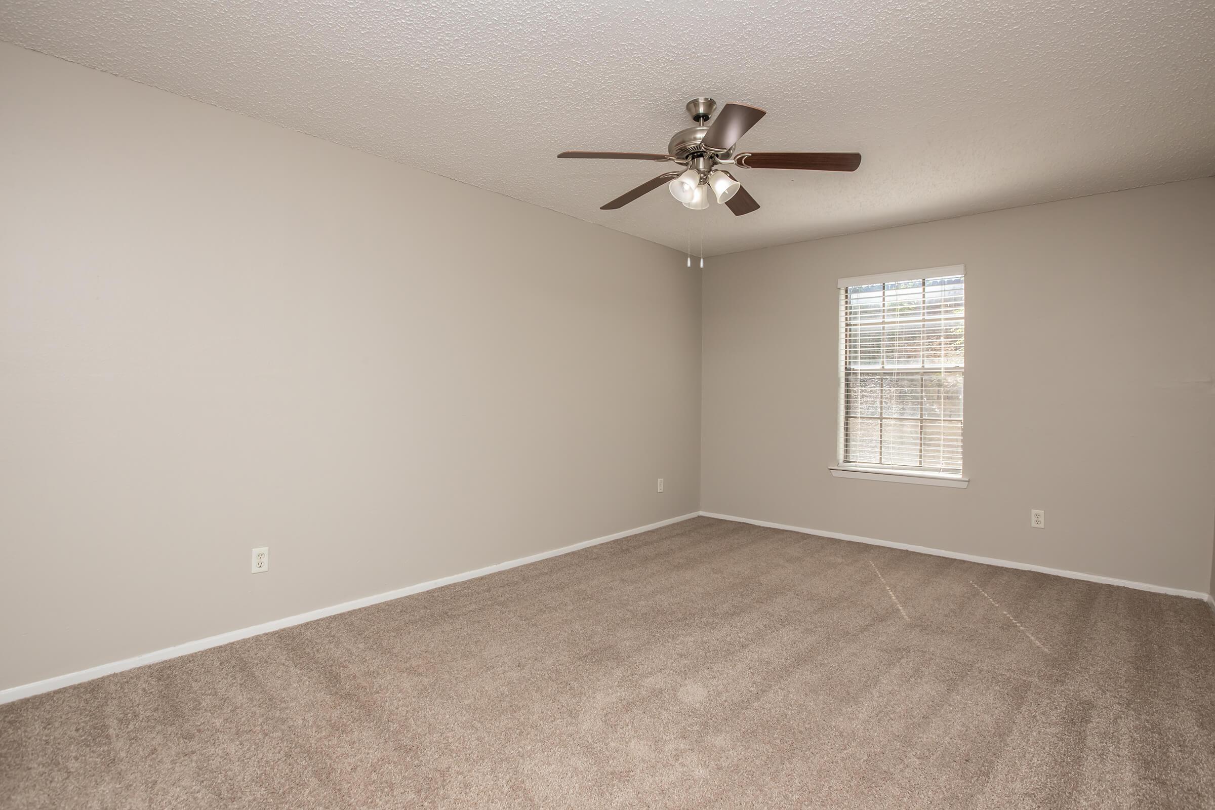 A spacious, empty room with light beige walls and carpet. It features a ceiling fan with three lights and a window with blinds that allows natural light to enter. The room is well-lit and clean, offering a simple and neutral aesthetic.