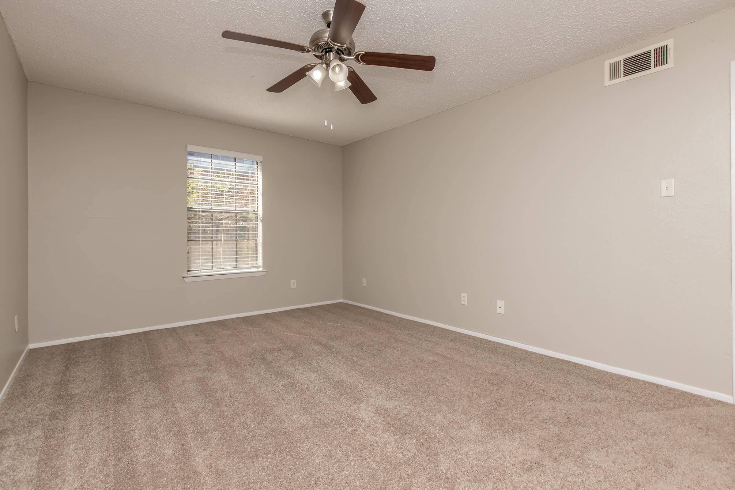 An empty room with light beige carpet and a ceiling fan. The walls are painted a soft gray, and there is a window with blinds that allows natural light to enter. The room appears spacious and unfurnished, with minimal decorations.