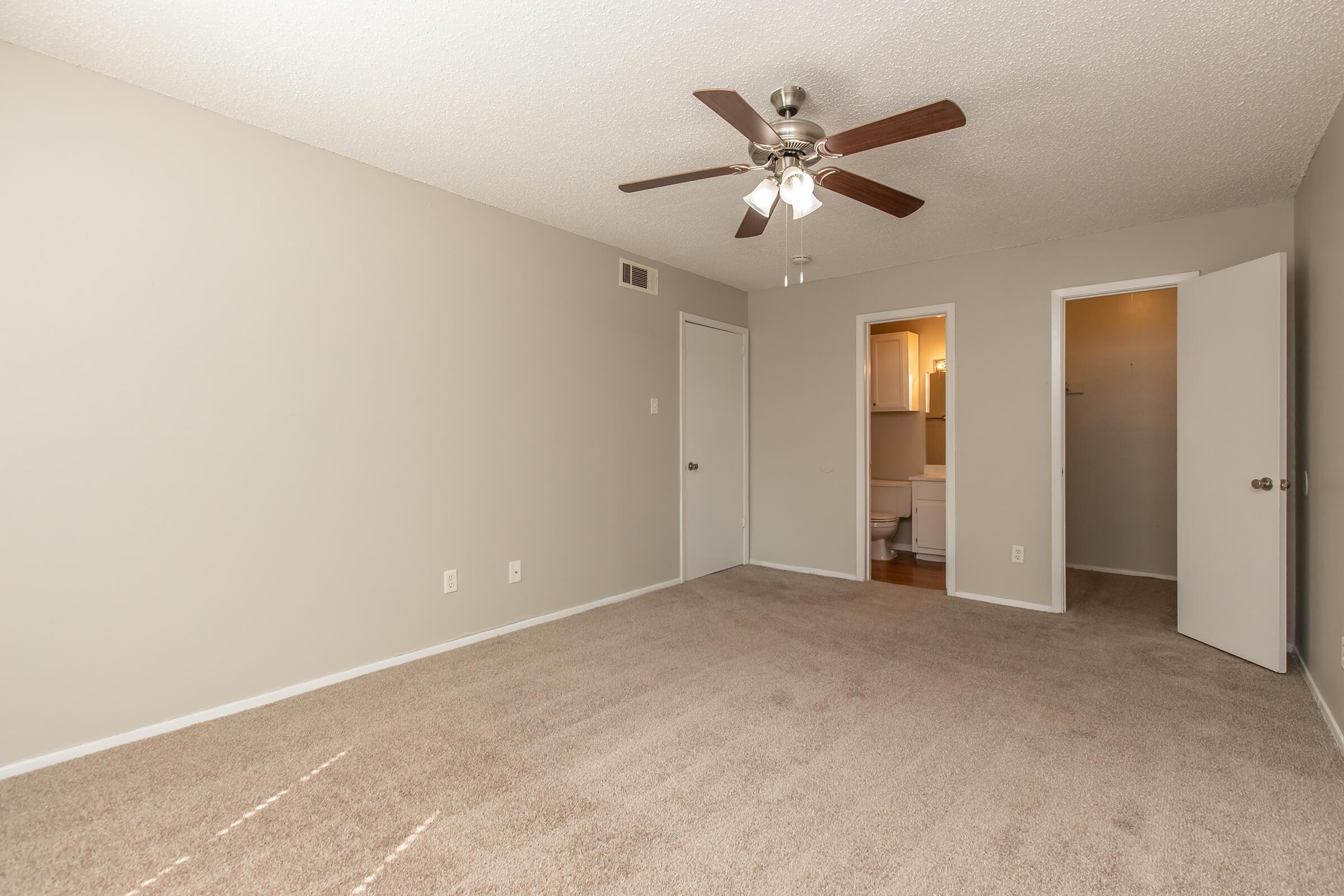 Empty room with light beige carpet and light gray walls. A ceiling fan is installed, and there are two doors leading to adjacent spaces. One door is partially open, revealing a bathroom area. Natural light filters in, creating a bright and spacious atmosphere.