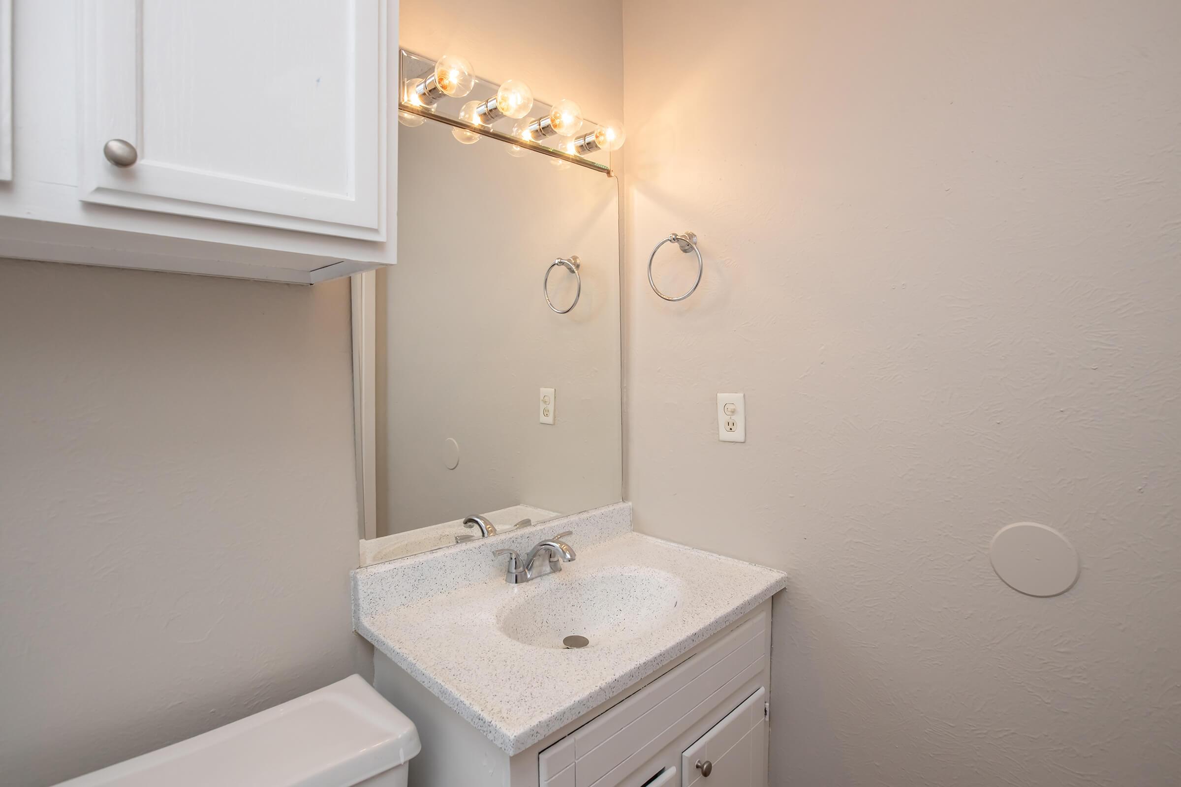 A small bathroom scene featuring a white vanity with a sink, a light fixture above the mirror, and a towel holder on the wall. The walls are a neutral color, and there is a white cabinet and a toilet visible in the corner. The setup is clean and minimalist, emphasizing functionality.
