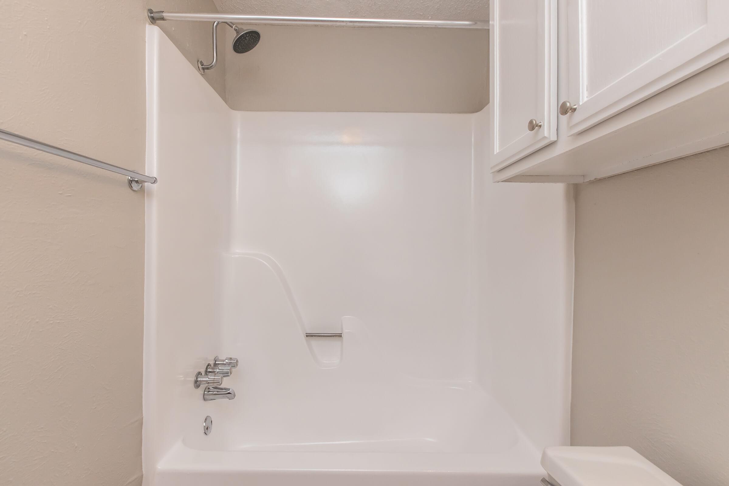 A clean, minimalist bathroom featuring a white bathtub with a showerhead above. The walls are plain and light-colored, and there are white cabinets mounted above. The overall look is bright and uncluttered, emphasizing simplicity and functionality.