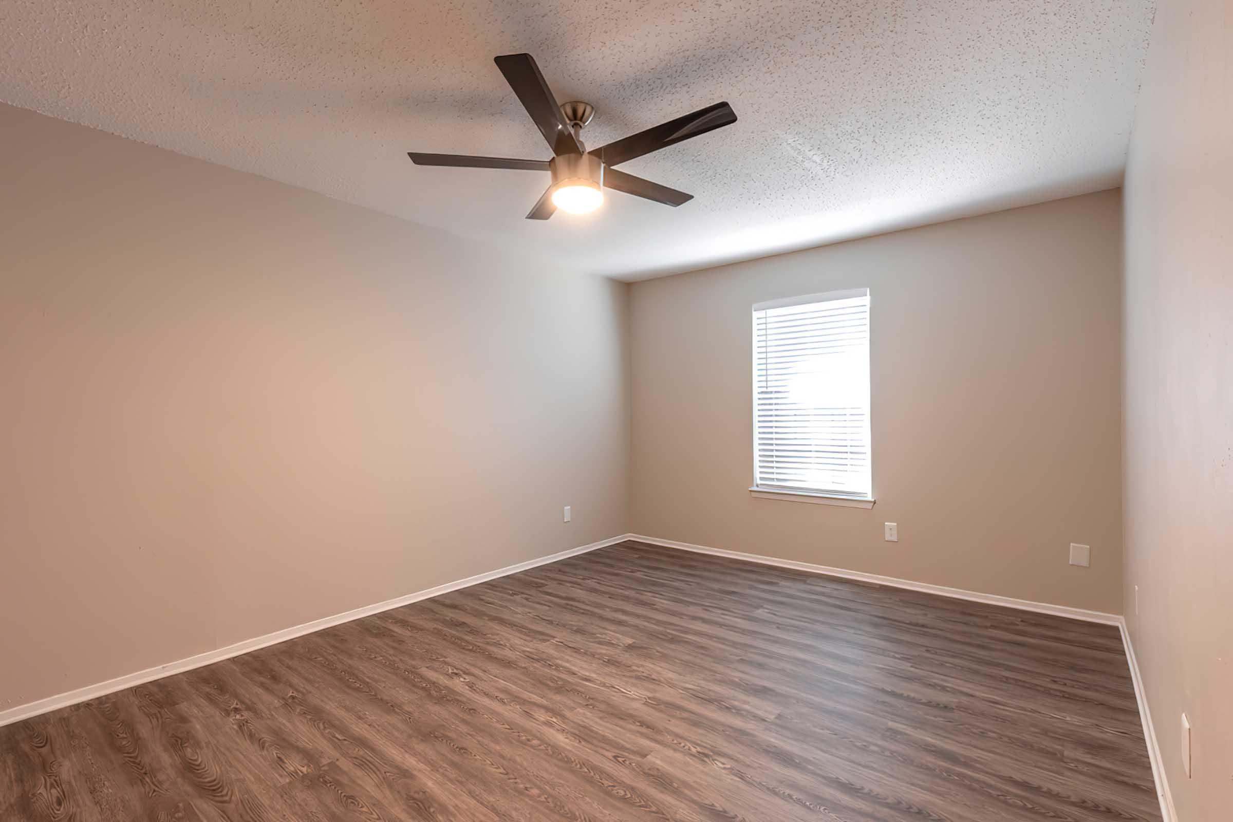 A spacious, empty room with light brown walls and a light wood floor. There’s a ceiling fan with a light fixture, and a window with white blinds letting in natural light. The room has a minimalist design, ideal for various uses.