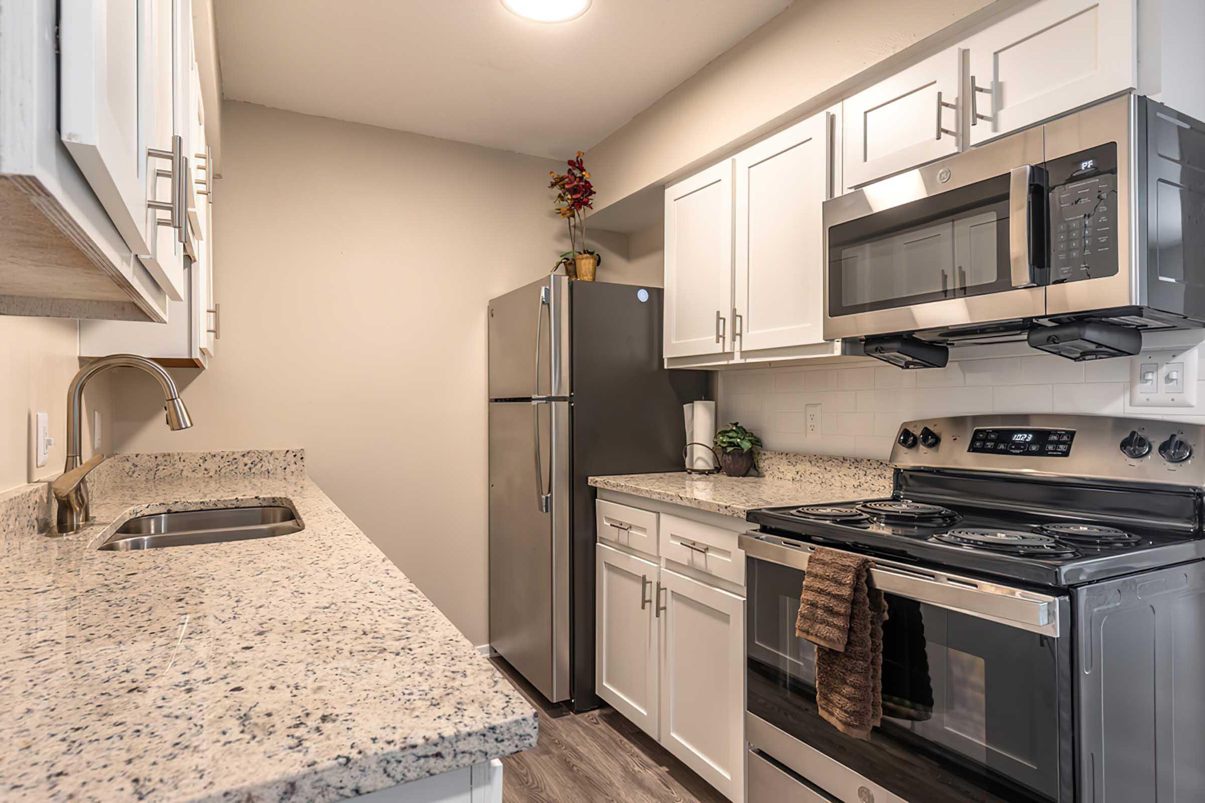 Modern kitchen featuring white cabinetry, granite countertops, stainless steel appliances including a refrigerator and microwave, and a stove with a backsplash. The space is well-lit with a subtle decorative plant on the counter.
