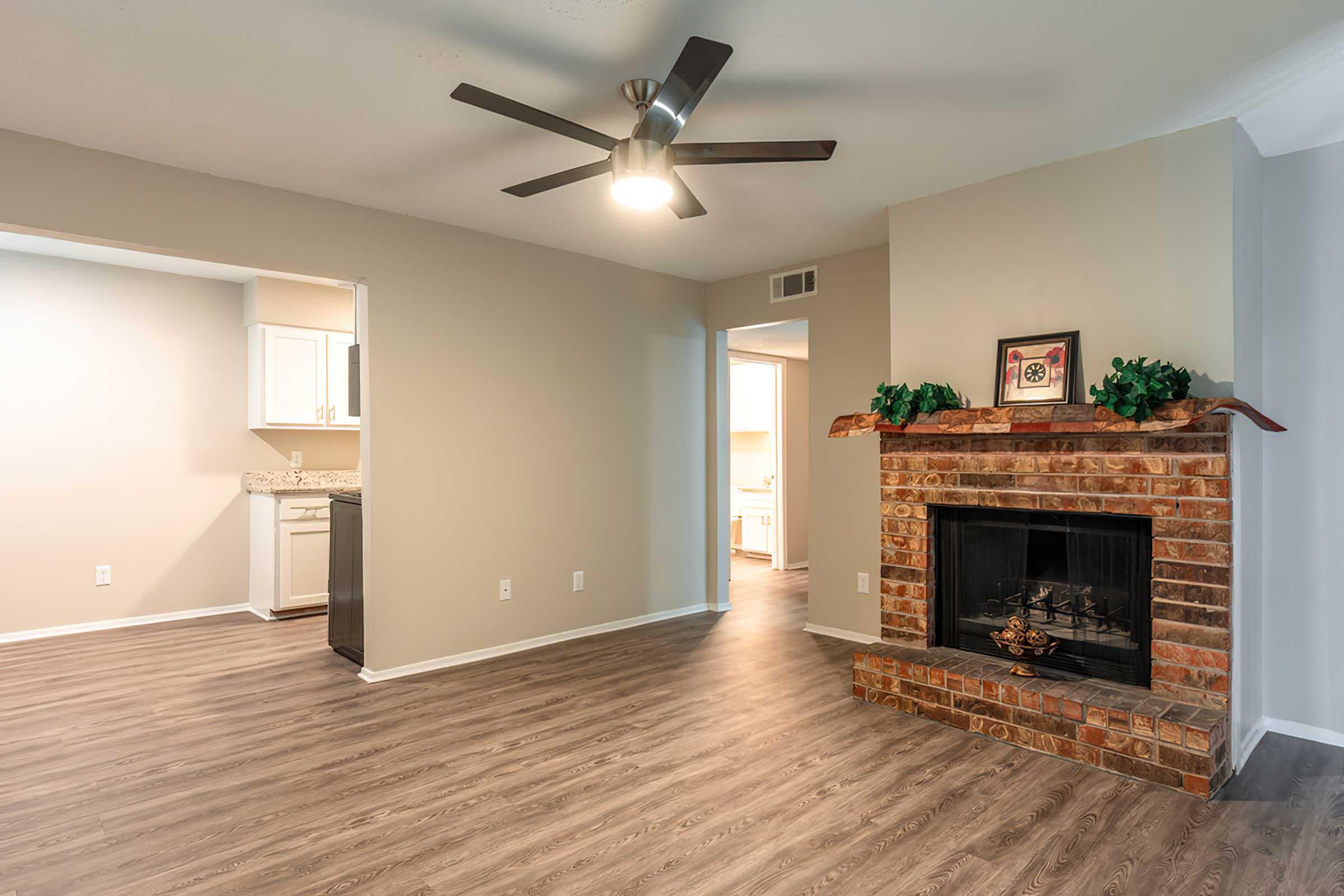 Spacious living room featuring light-colored walls, a modern ceiling fan, and a cozy brick fireplace with decorations. To the left, a doorway leads to a kitchen with white cabinets, while a hallway can be seen in the background, adding warmth and charm to the open floor plan.