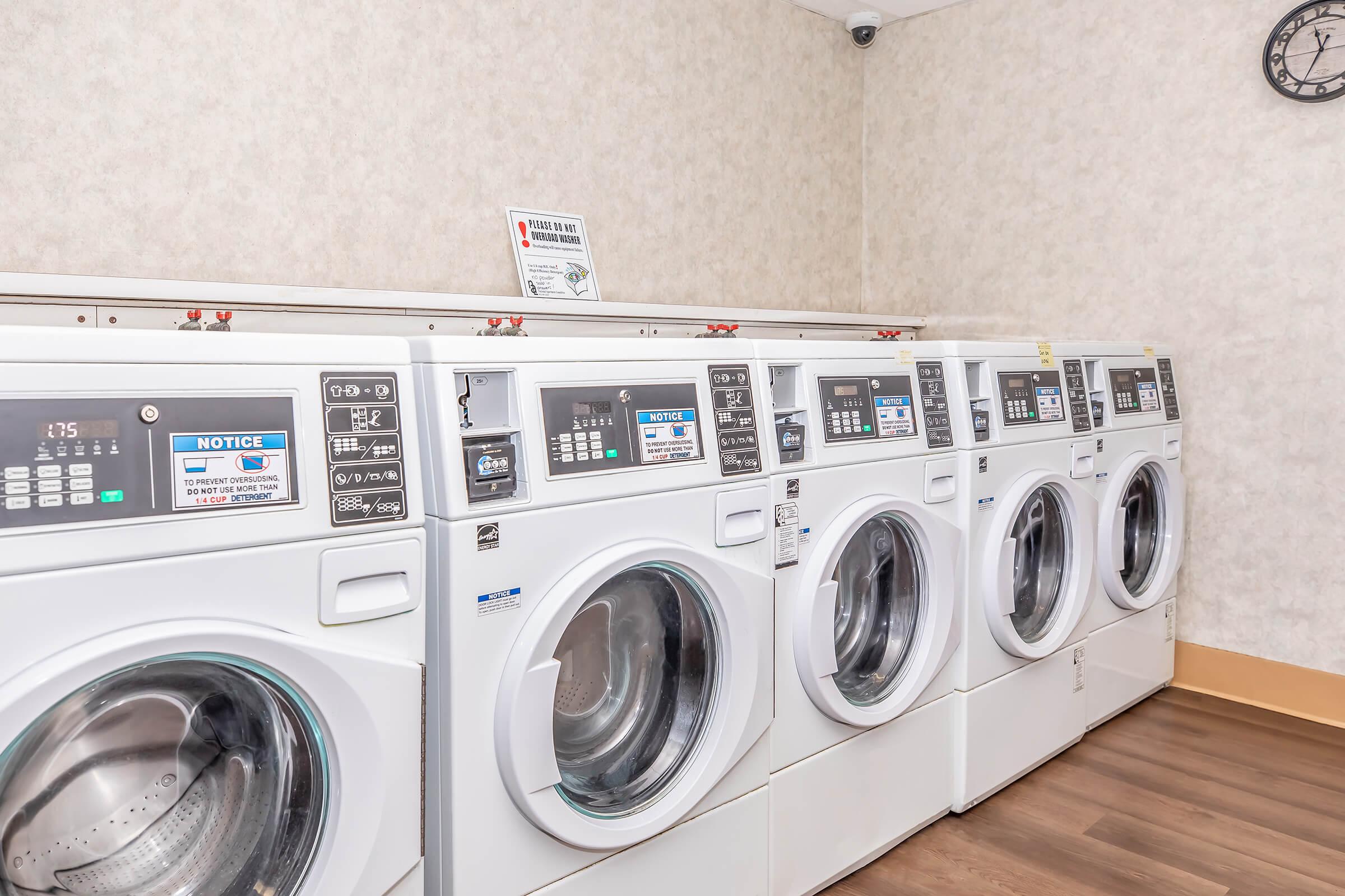 A row of modern, front-loading washing machines in a laundry room. They feature digital controls and clearly labeled settings. The floor is wooden, and the walls have a light-colored wallpaper. A notice sign is visible above the machines, and a clock is mounted on the wall in the background.