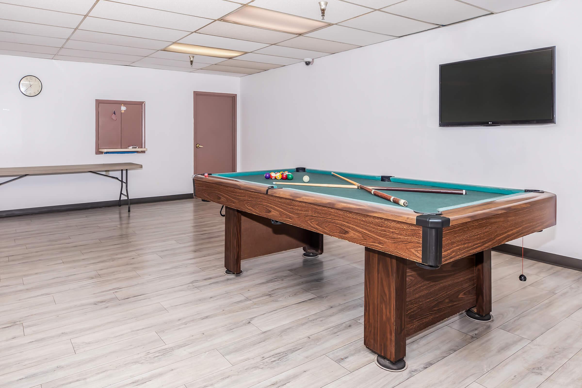 A clean, well-lit room featuring a wooden pool table with a green felt surface, cues, and colored balls. There is a wall-mounted television and a clock on the wall, along with a folding table in the background. The flooring is light-colored with a modern design.