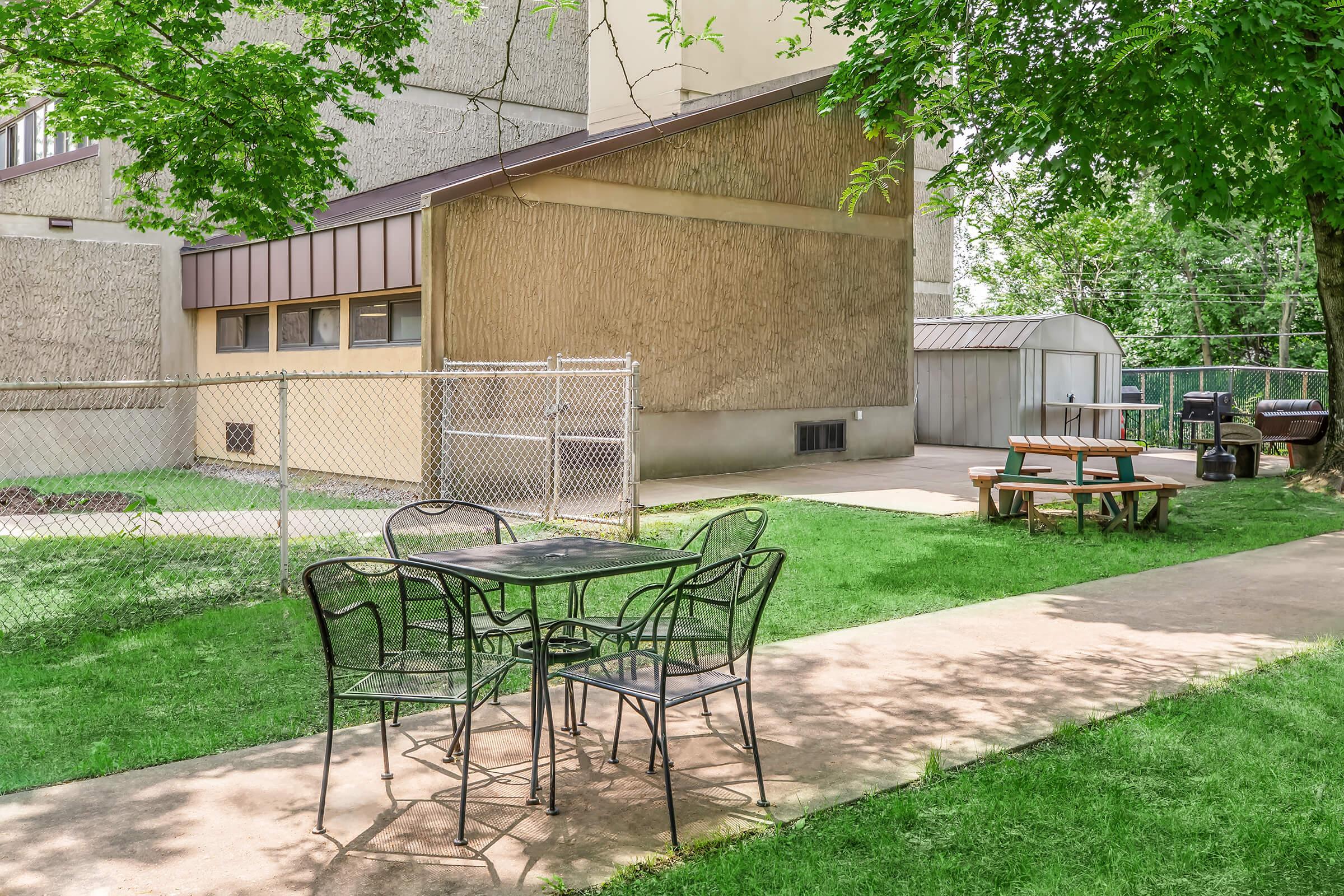A fenced outdoor area featuring a picnic table and a small black table with four chairs. The background includes a building with textured walls and a shed. Lush green grass surrounds the seating area, creating a pleasant space for outdoor relaxation or gatherings.