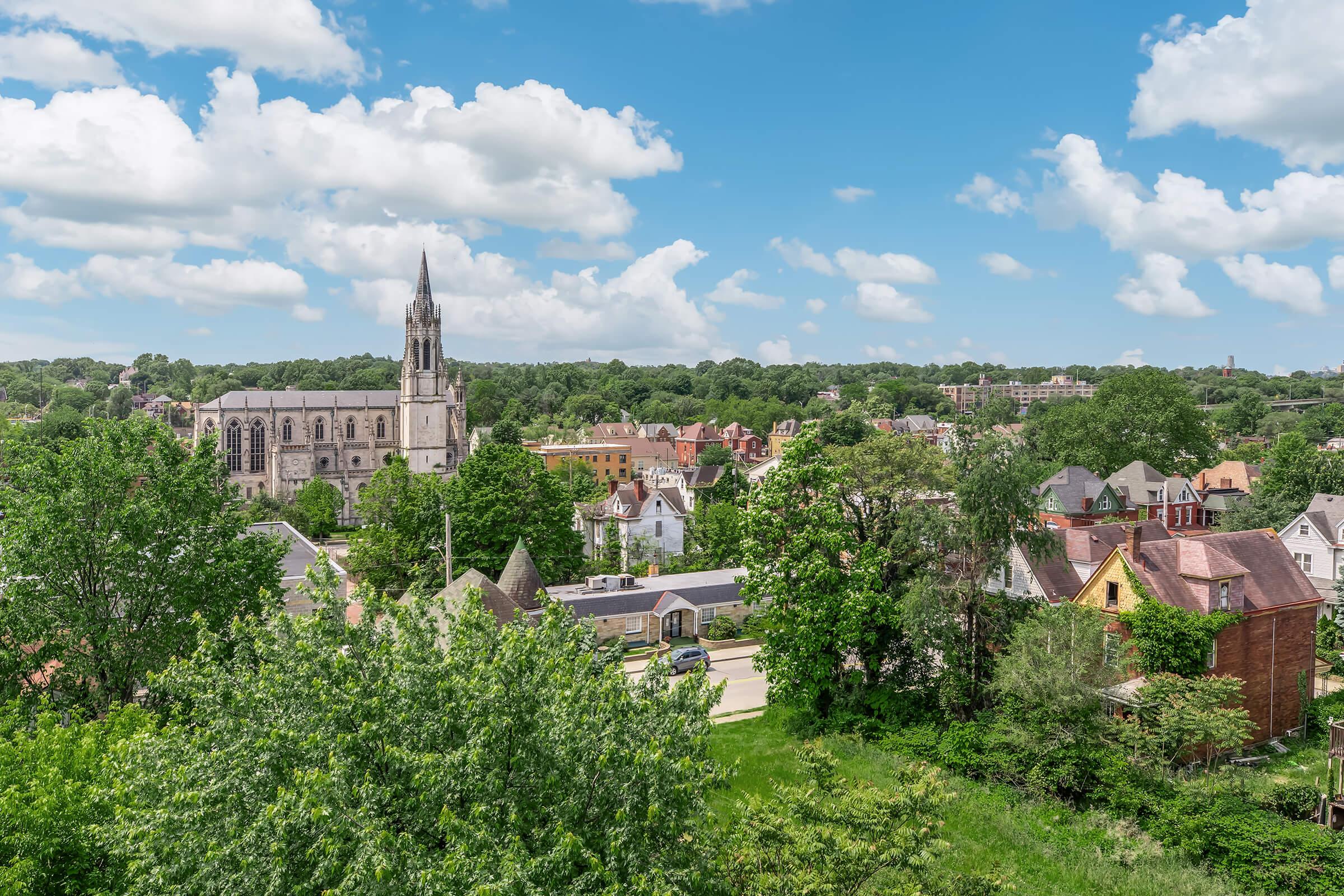A scenic view of a town featuring a historic church with a tall steeple, surrounded by greenery and residential buildings. The sky is bright blue with fluffy white clouds, and the landscape includes trees and well-maintained homes, showcasing a peaceful community vibe.