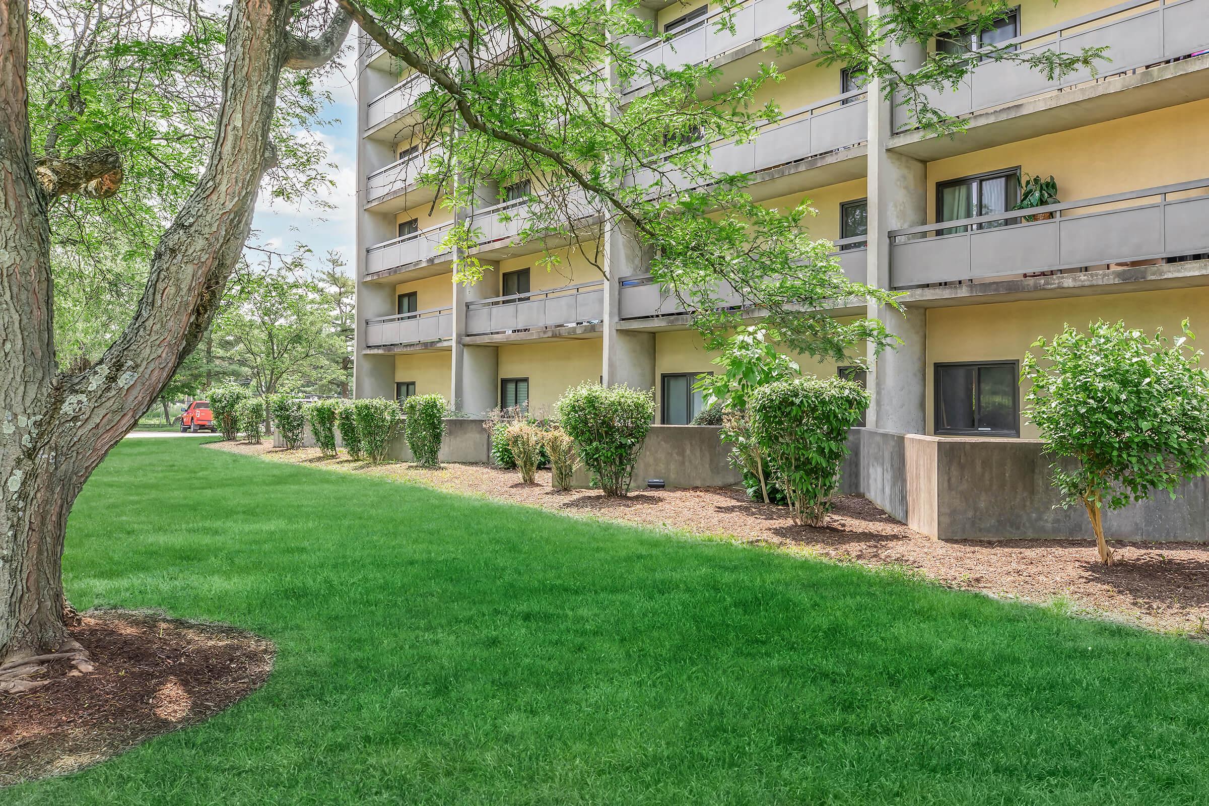 A grassy area beside a multi-story building, featuring yellow walls and several balconies. Trees provide shade, and neatly trimmed shrubs line the pathway. There's a hint of outdoor seating visible on one balcony. The scene conveys a serene, well-maintained environment.