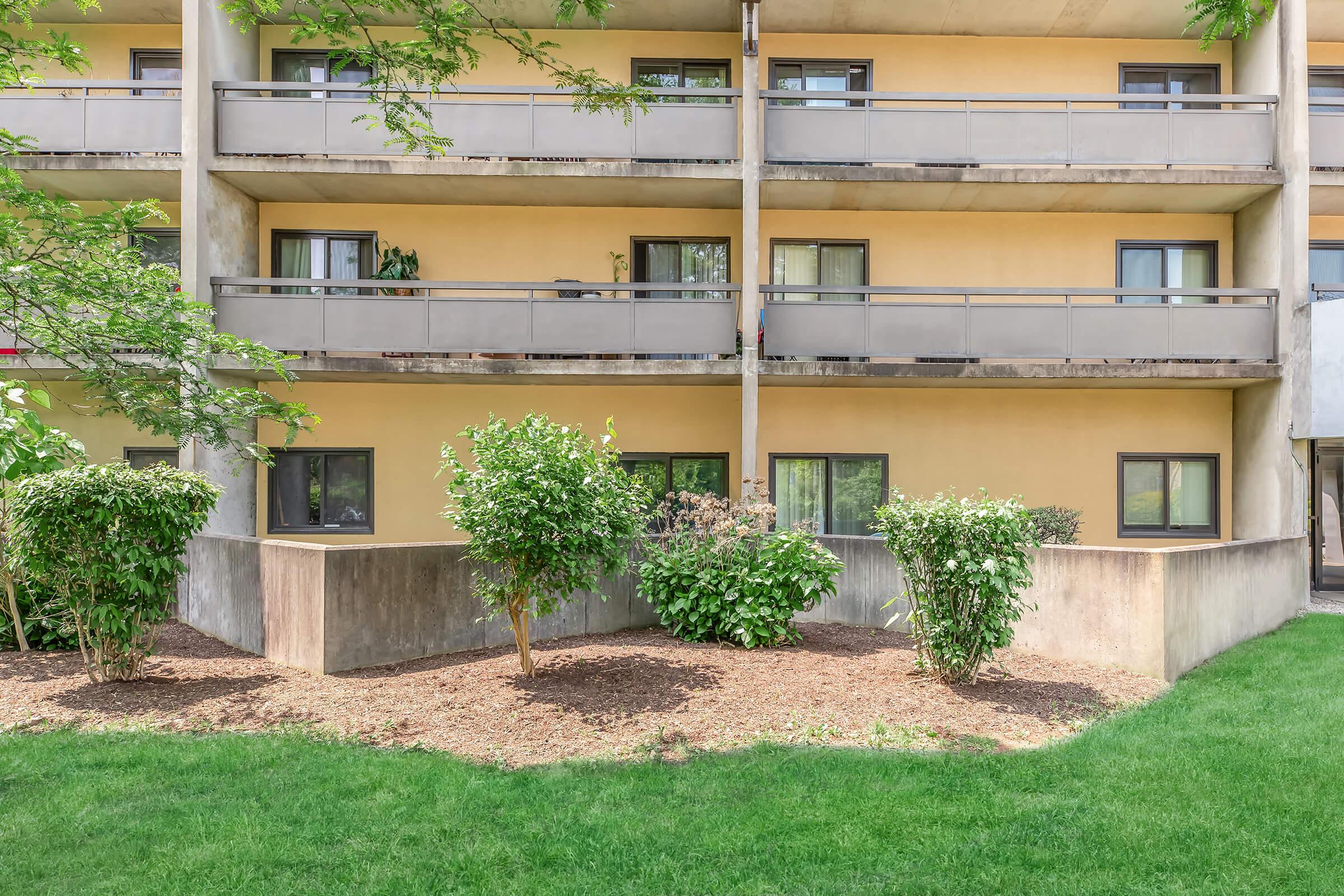 A view of a modern multi-story building featuring balconies with plants and small shrubs in the foreground. The ground is covered in mulch, and there is a well-maintained lawn, creating a tidy and inviting outdoor space. The architecture combines concrete and a bright yellow facade.