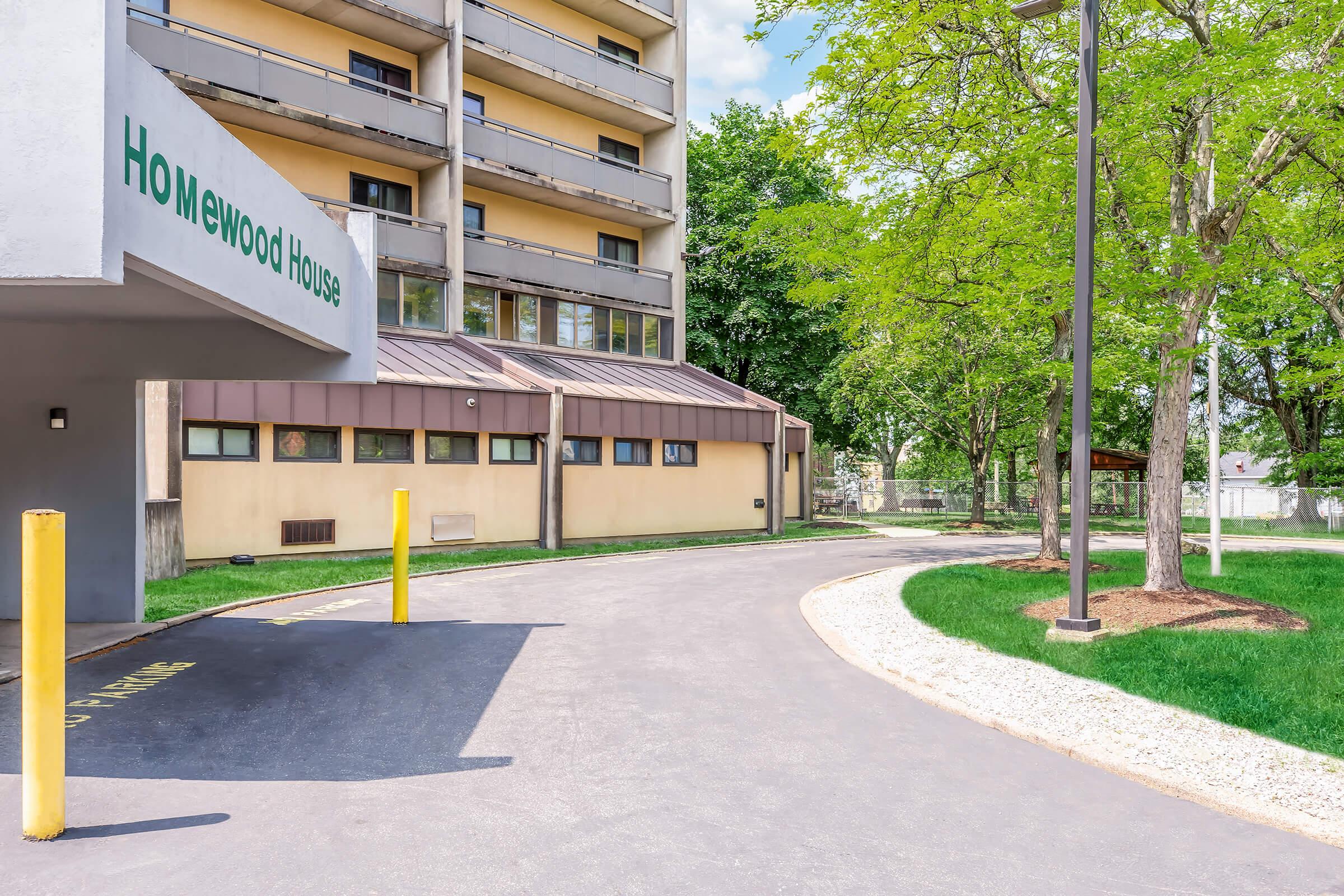 View of Homewood House building, featuring a sloped driveway with yellow bollards and landscaped green areas. The structure has multiple floors and large windows, surrounded by trees and a well-maintained pathway. The sky is clear, indicating a bright day.