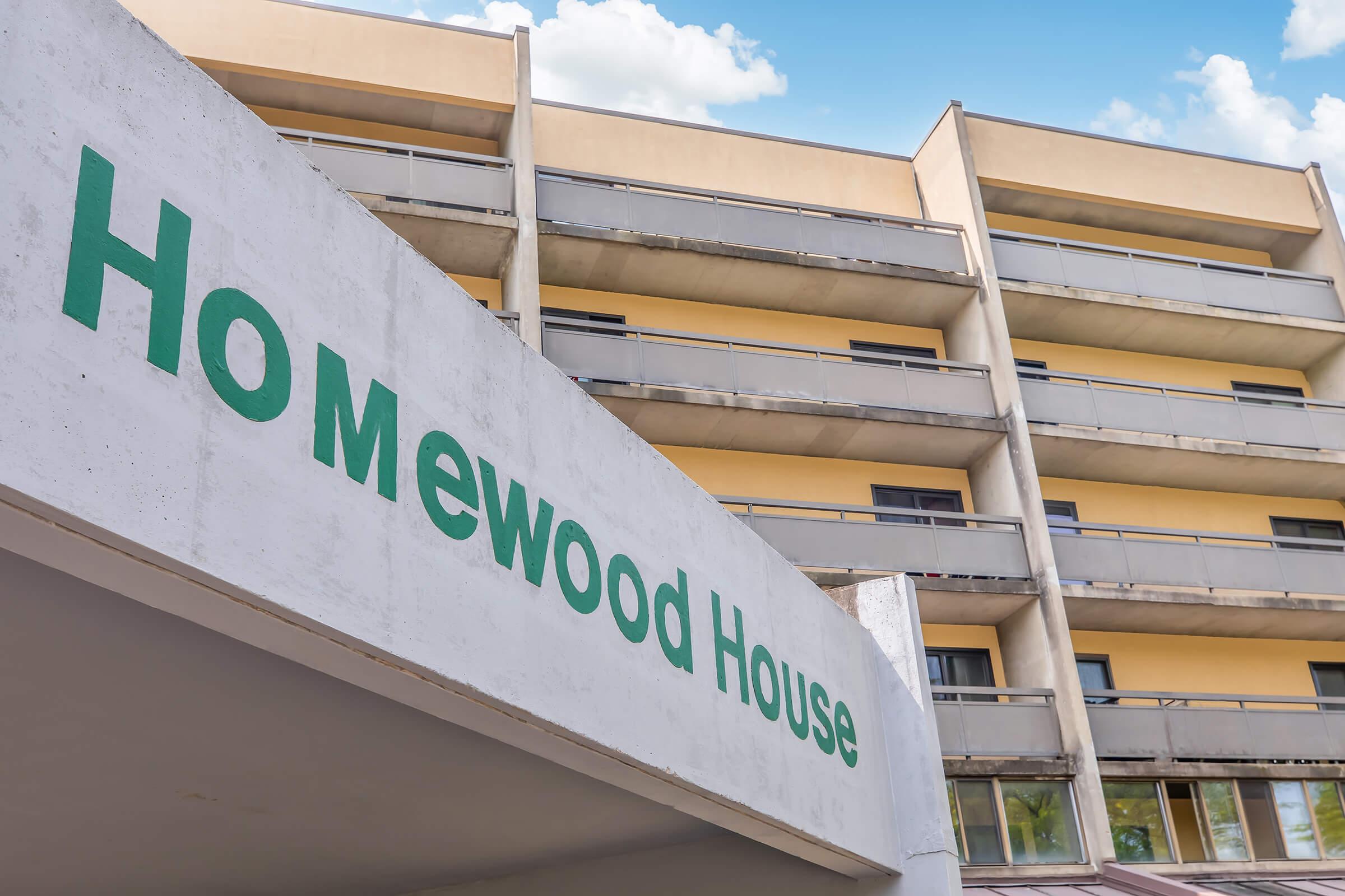 Sign with the text "Homewood House" prominently displayed in green on a white wall, in front of a multi-story building with balconies. The building features a modern design with light-colored walls and a clear blue sky overhead.
