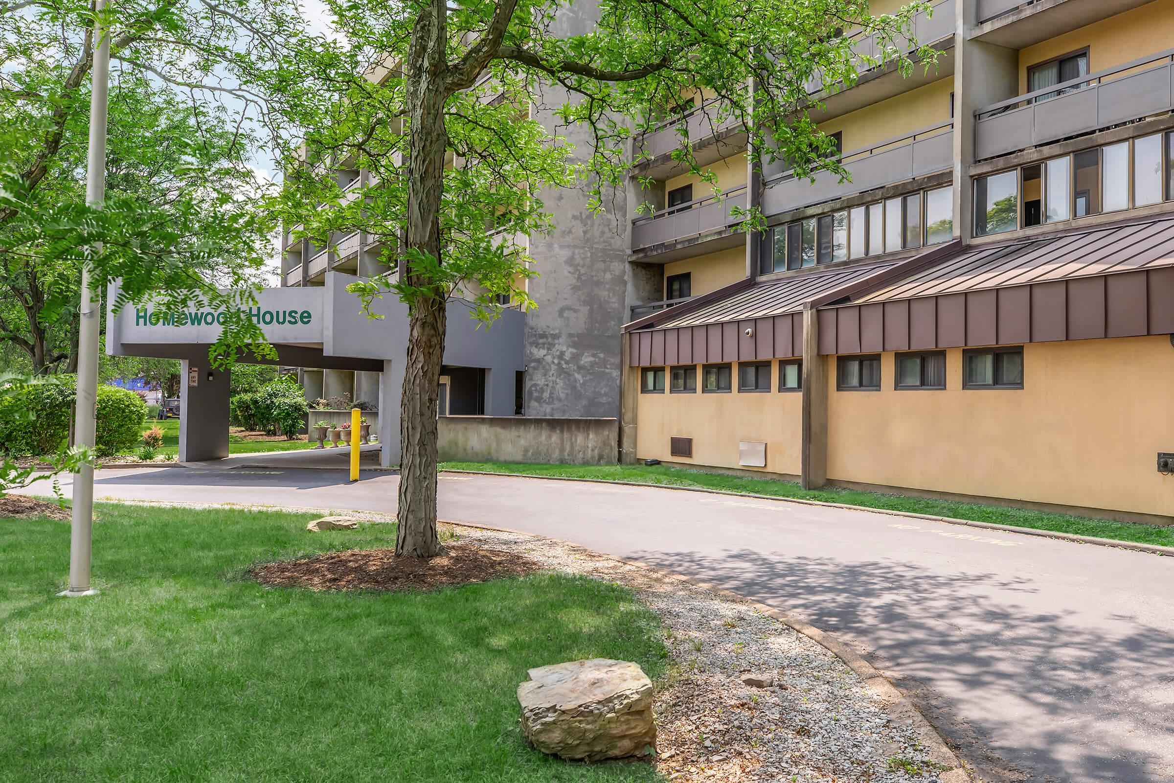 A view of Homewood House, featuring a modern building with a gray facade and distinct architectural elements. The area is landscaped with green grass and trees, and a paved pathway leads to the entrance. The surroundings are well-maintained, showcasing a blend of natural and urban design.