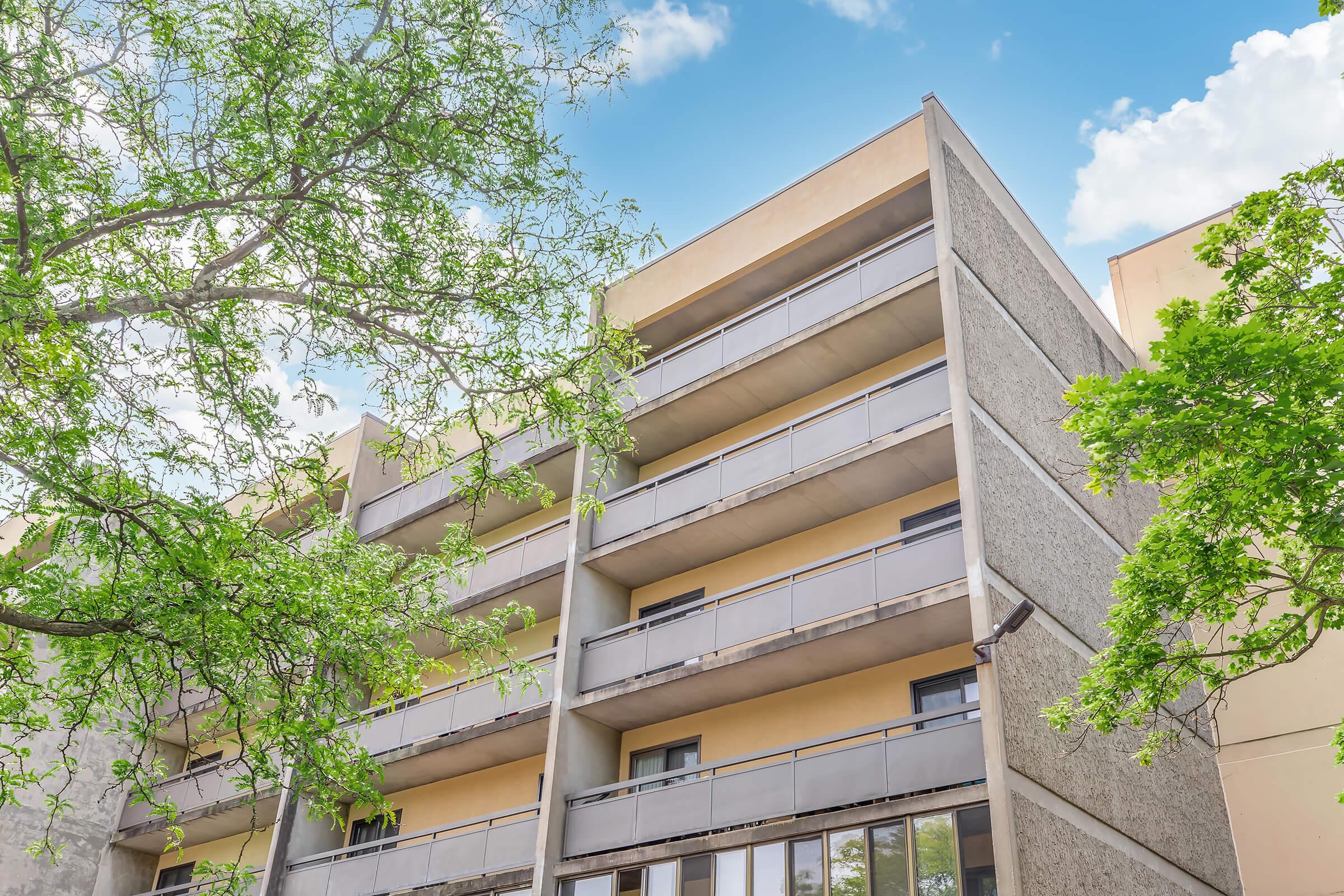 A modern multi-story building with balconies, set against a bright blue sky with a few clouds. Green leaves from nearby trees frame the image, adding a touch of nature to the urban scene. The building's façade features a mix of concrete and yellow-colored walls.