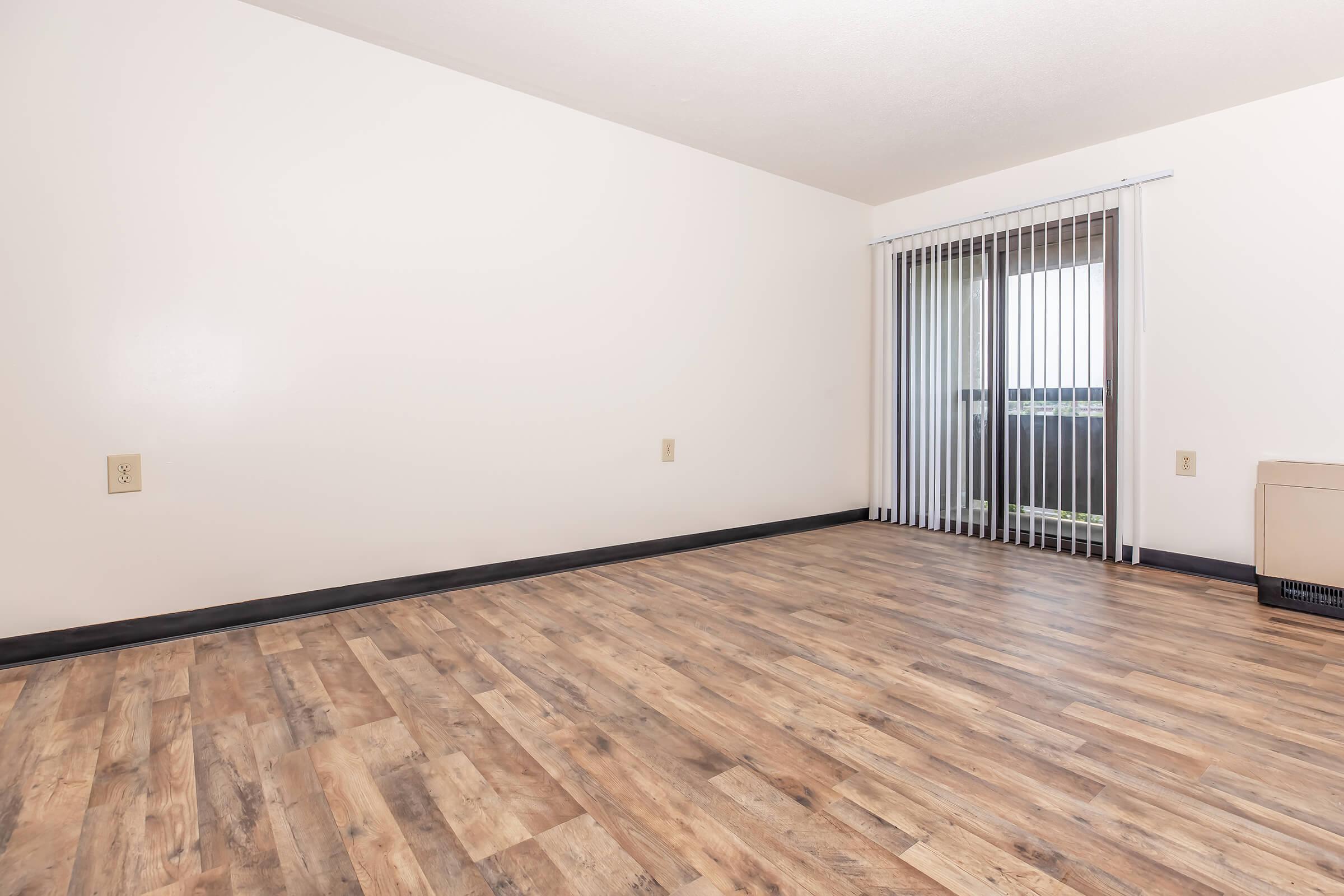 Empty interior of a room featuring light-colored walls, a large window with vertical blinds, and a floor made of wood-like laminate. The space has a clean, minimalist appearance with no furniture, creating a bright and airy atmosphere.