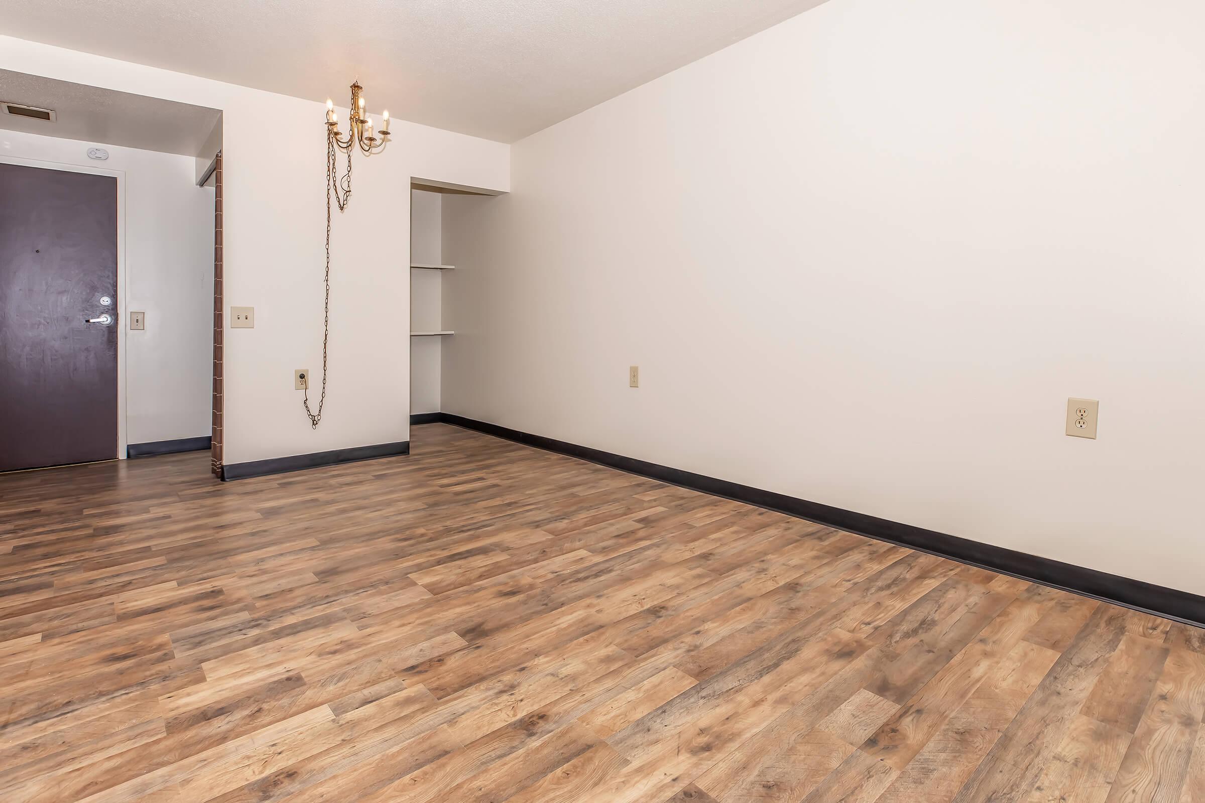 A spacious, empty room featuring light-colored walls and a wooden floor. A small chandelier hangs from the ceiling, and there is a doorway on the left leading to another area. A shelf is built into the wall on the right, providing additional storage or display space.