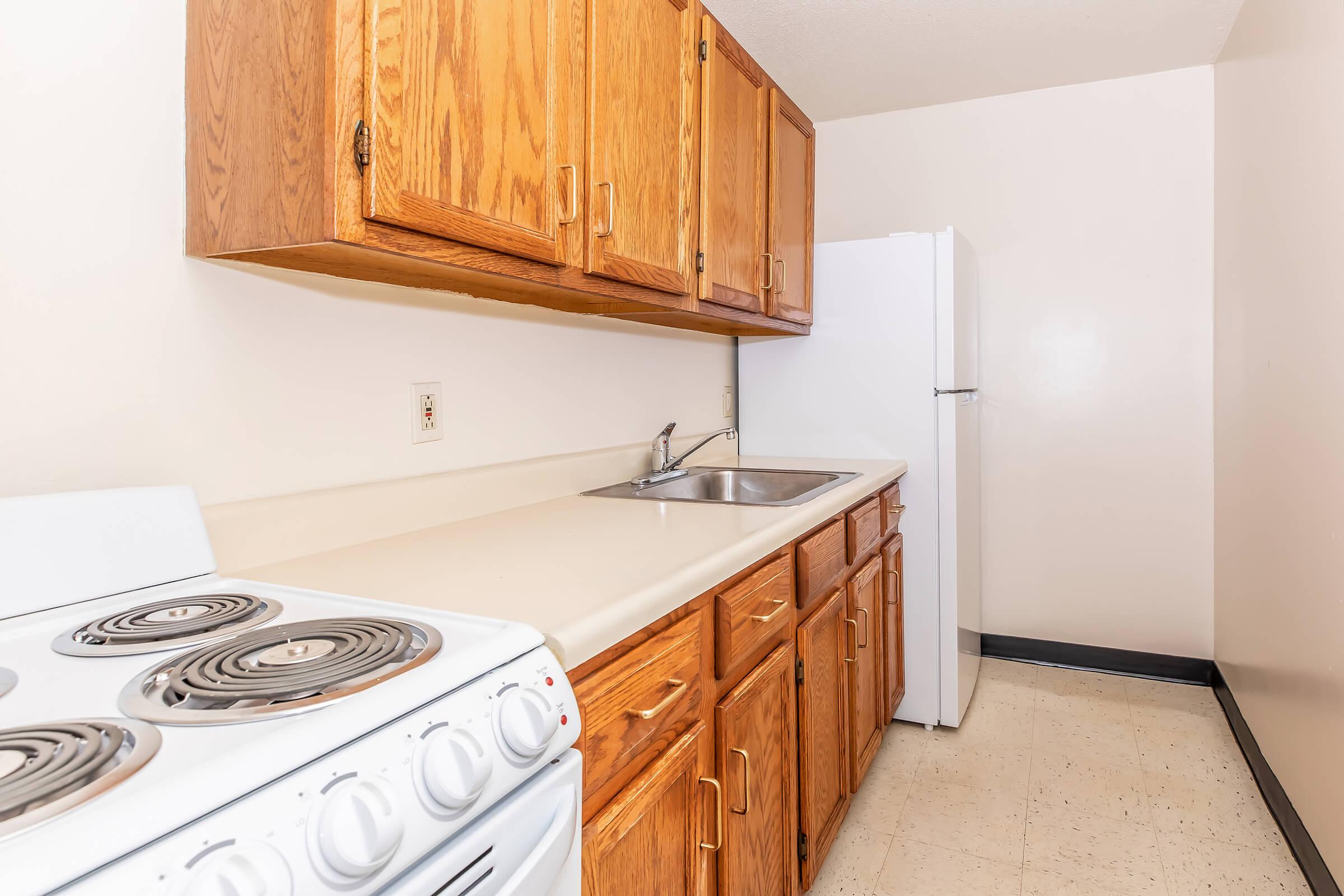 A kitchen featuring light-colored countertops, wooden cabinets, a white refrigerator, and a white stove with four burners. The walls are painted a neutral color, and the floor is tiled in a light shade. The overall space appears clean and functional.