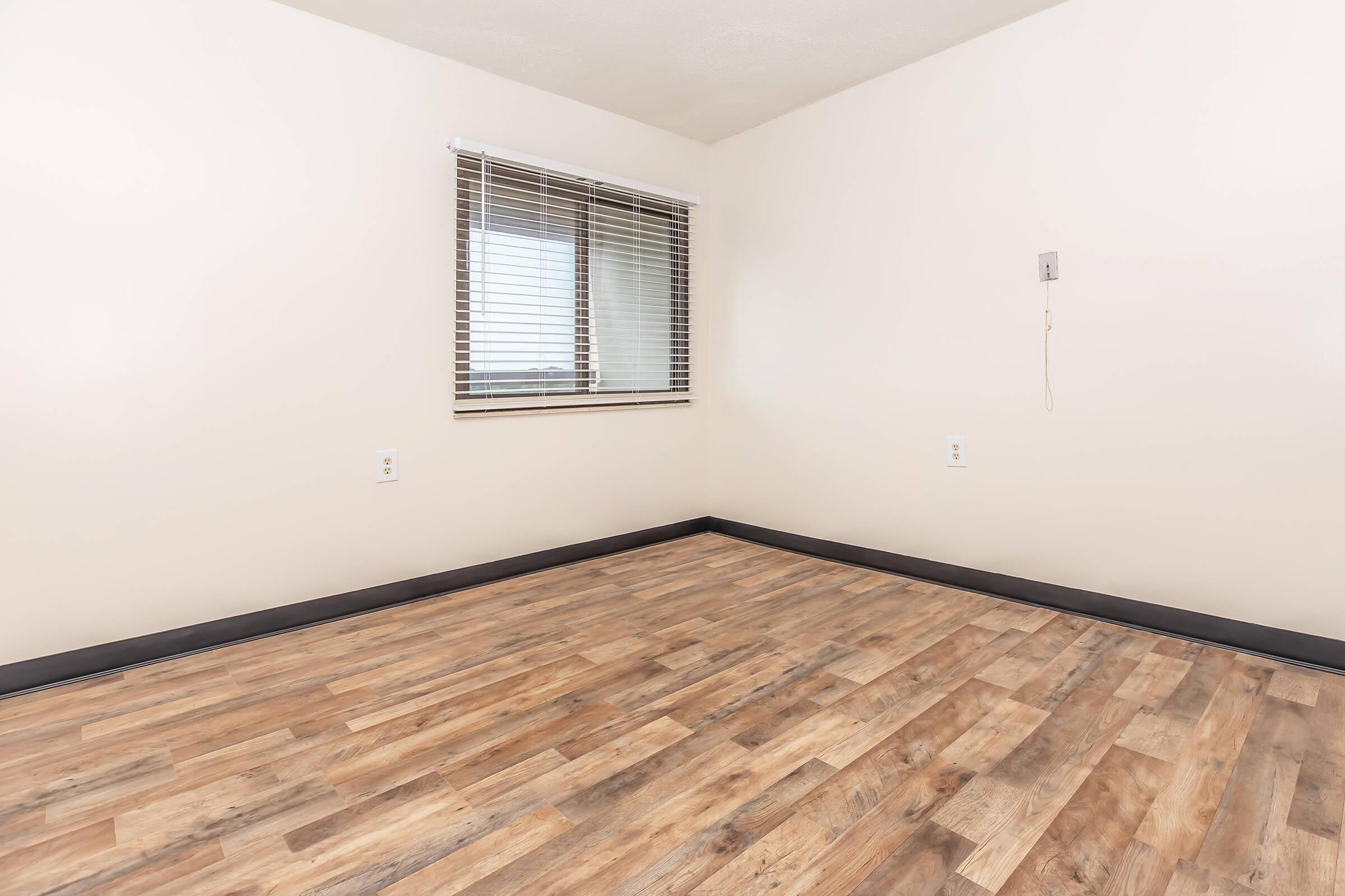 A spacious, empty room with light-colored walls and a large window featuring blinds. The floor is covered in wood-like laminate, and there are electrical outlets on the wall. A single light switch is visible, and the room has a clean, minimalistic aesthetic, making it suitable for various uses.