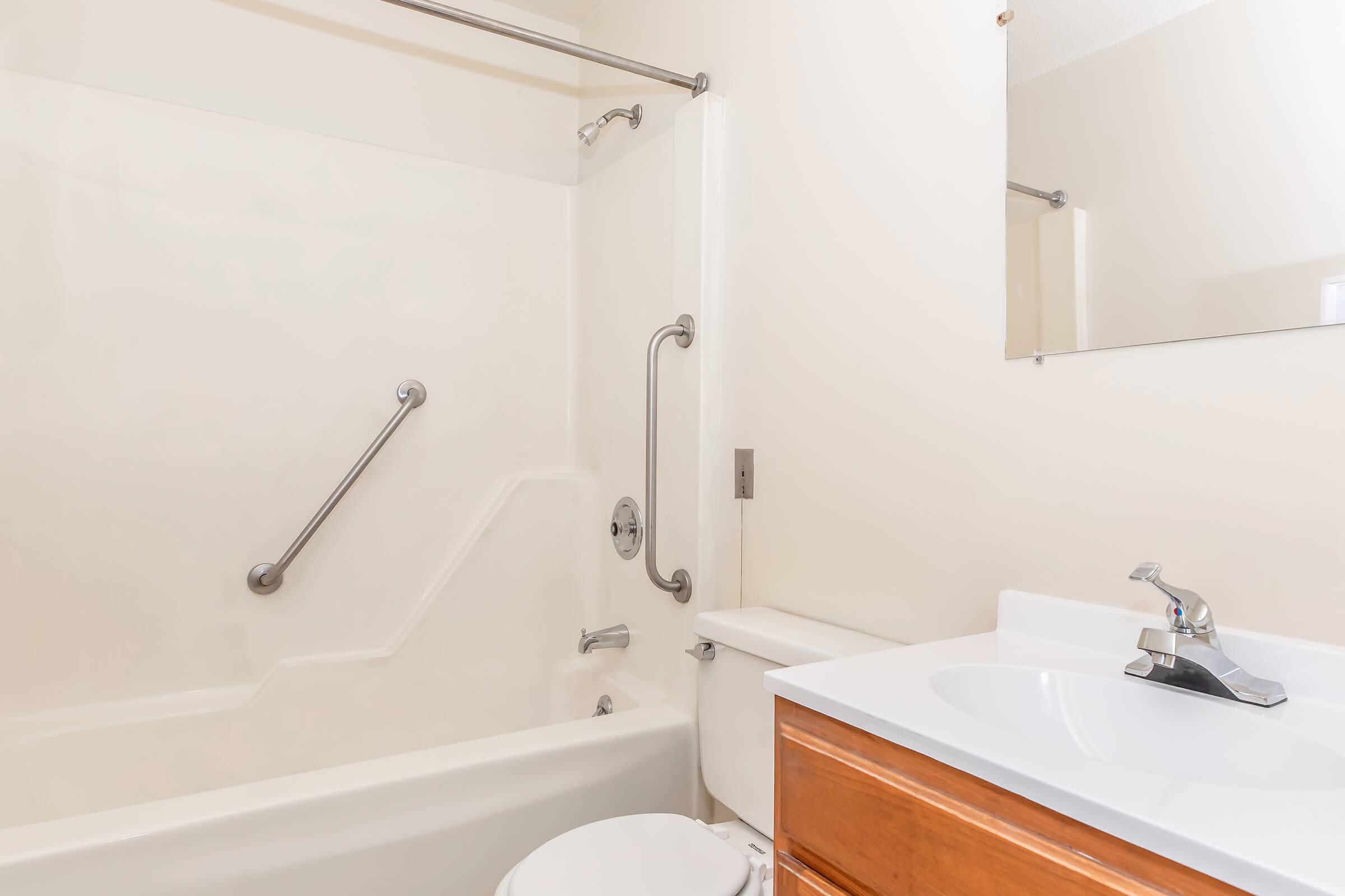 A clean, well-lit bathroom featuring a bathtub with a shower, grab bars for safety, a simple white sink with a wooden cabinet below, and a mirror above the sink. The walls are painted in a light color, contributing to a spacious and airy feel.