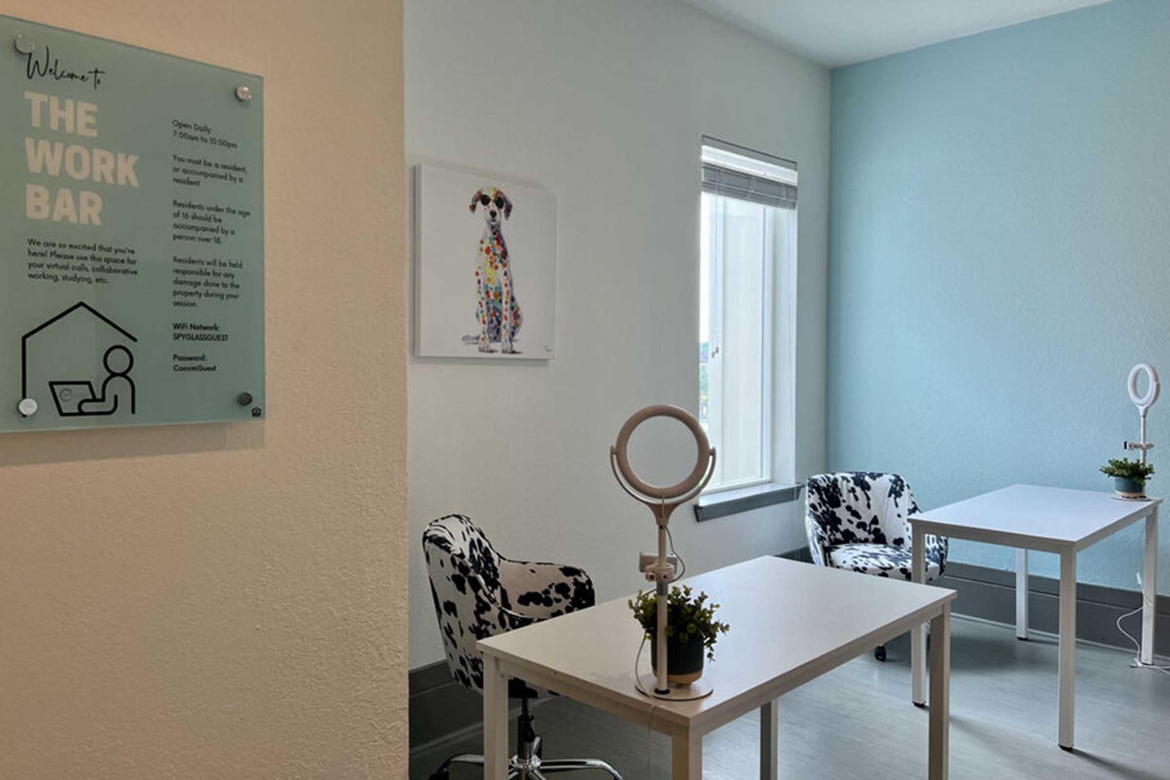 A bright and modern workspace featuring two white desks with chairs that have a cow-print design. There are small potted plants on the desks and a large window allowing natural light. A sign on the wall reads "Welcome to THE WORK BAR" with additional information about the space.