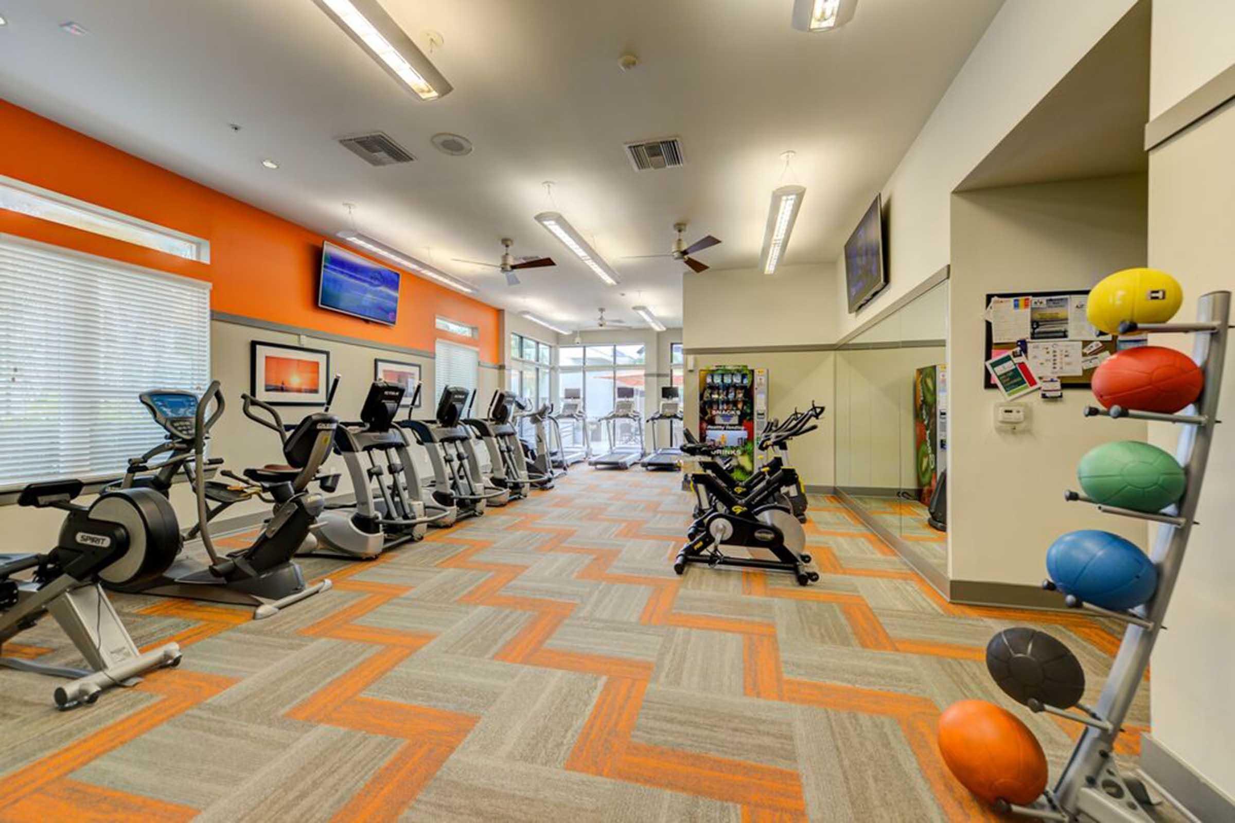 A bright and modern gym space featuring exercise equipment such as stationary bikes, treadmills, and a multi-functional workout machine. The room has large windows providing natural light, orange accent walls, and a colorful rack of exercise balls, creating an inviting atmosphere for fitness activities.