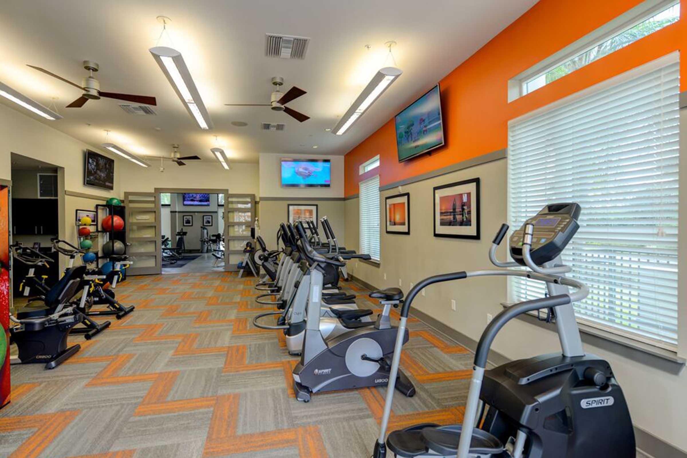 A modern gym interior featuring exercise equipment like treadmills and ellipticals, bright orange accent walls, and large windows allowing natural light. There are multiple screens displaying fitness content, and the space is neatly organized with a light gray and orange color scheme.