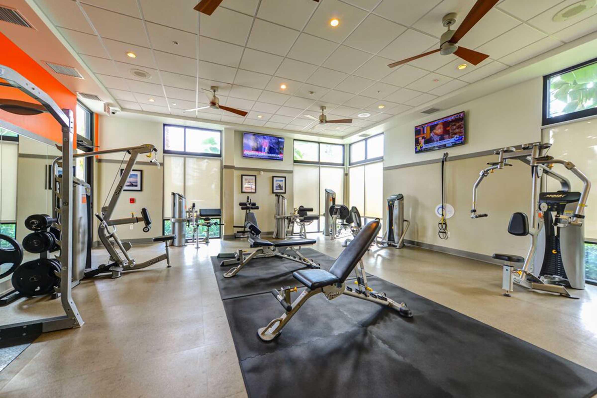 A clean, modern gym space featuring various workout equipment including weight machines, benches, and free weights. Large windows provide natural light, and two televisions are mounted on the walls, enhancing the workout environment. The flooring is a mix of rubber and tile.