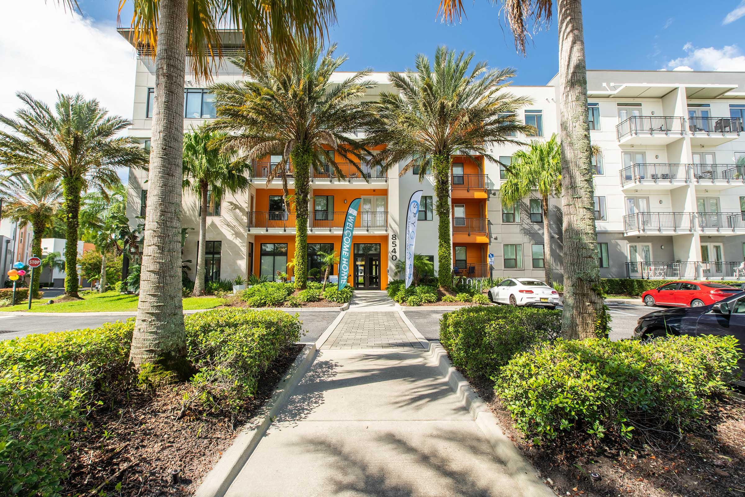 A modern apartment building with palm trees and lush landscaping. The entrance features a welcoming design with orange accents and large windows. A pathway leads from the street to the entrance, with parked cars visible in the foreground. Bright blue sky and sunlight enhance the cheerful atmosphere.