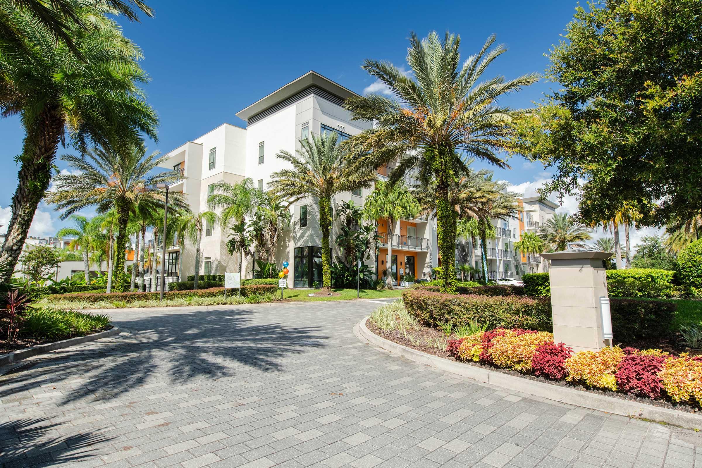 A modern building surrounded by lush palm trees and colorful landscaping. The entrance features large glass doors, and a winding stone pathway leads up to the entrance. Sunlight casts a warm glow over the scene, highlighting the vibrant greenery and well-maintained flowers.