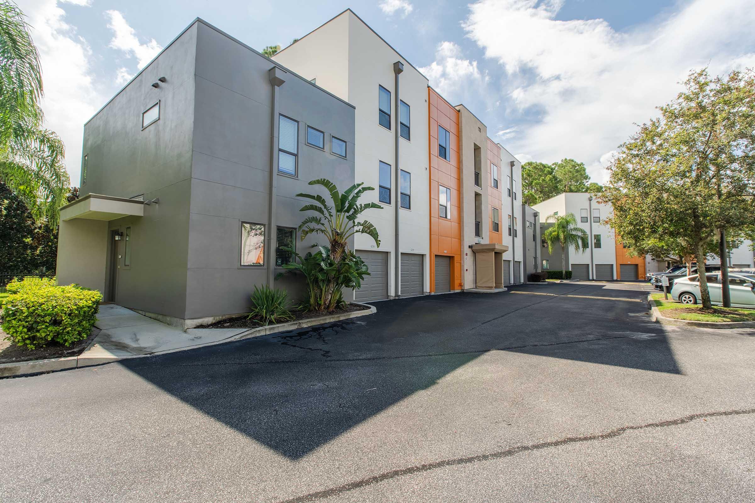 Two modern, multi-story residential buildings with a mix of grey and orange facades. The buildings are surrounded by landscaping with palm trees and shrubs. A paved driveway leads in front of the structures, providing access to parking spaces. The sky is partly cloudy, adding a bright ambiance to the scene.