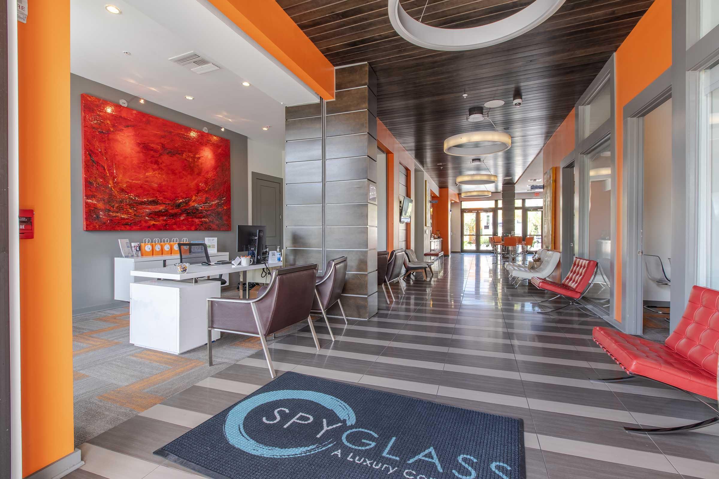 Modern reception area of a luxury apartment complex featuring a bright red abstract painting on the wall, sleek furniture, and a reception desk. The flooring is a mix of tiles, and there is a prominent blue welcome mat that reads "SPYGLASS." Large windows allow natural light to fill the space.