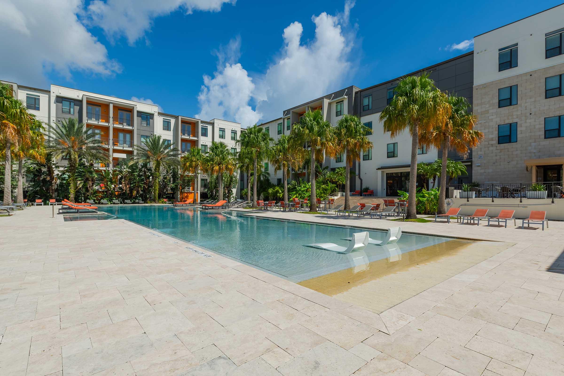A sunny hotel courtyard featuring a large swimming pool surrounded by palm trees and lounge chairs. Modern apartment buildings with balconies are visible in the background, under a blue sky with scattered clouds. The area is designed for relaxation and recreation.