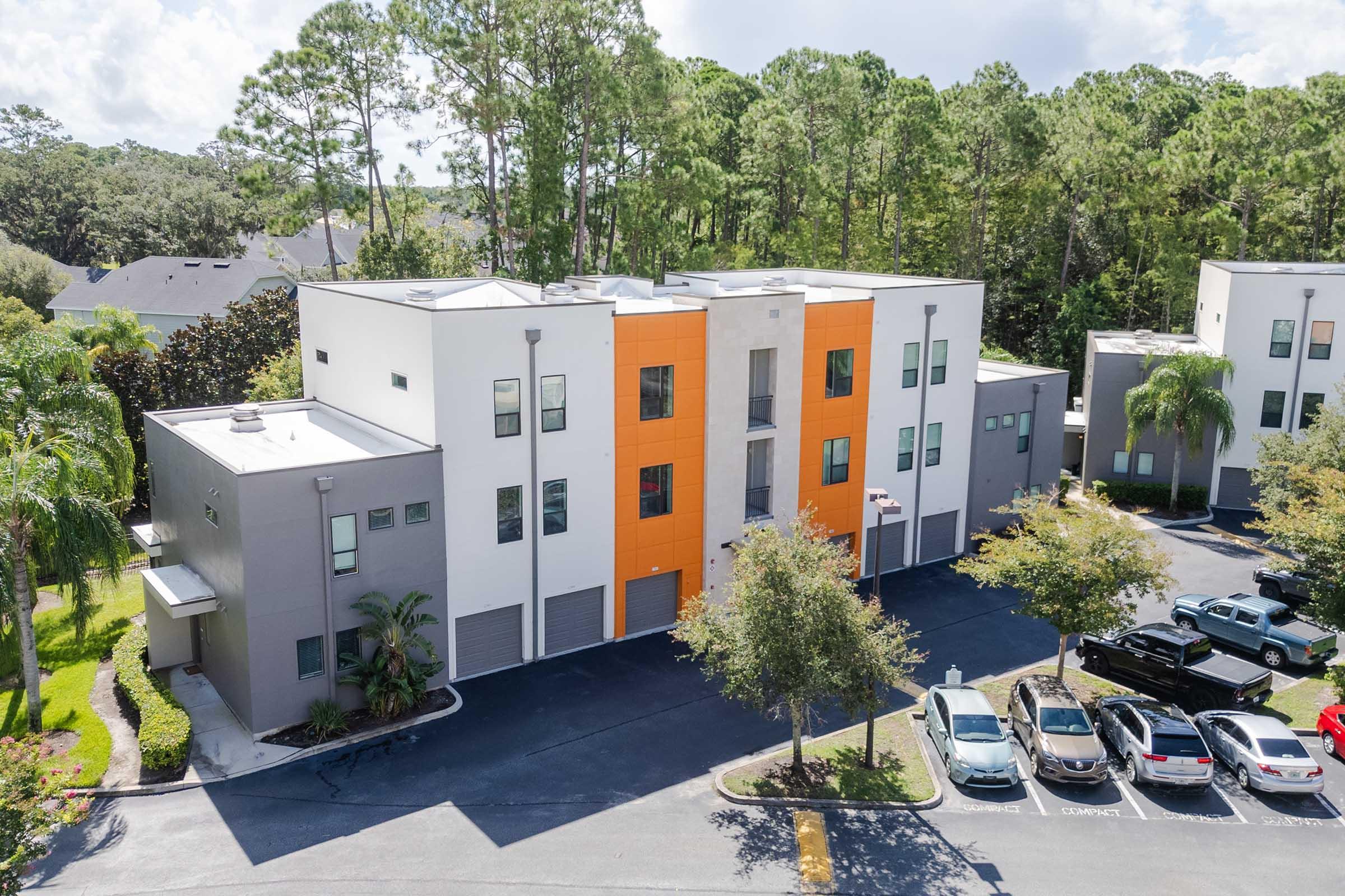 A modern, three-story apartment building surrounded by greenery. The building features a mix of white and orange paneling. In the foreground, several parked cars are visible along the driveway, with trees and landscaping enhancing the outdoor environment.
