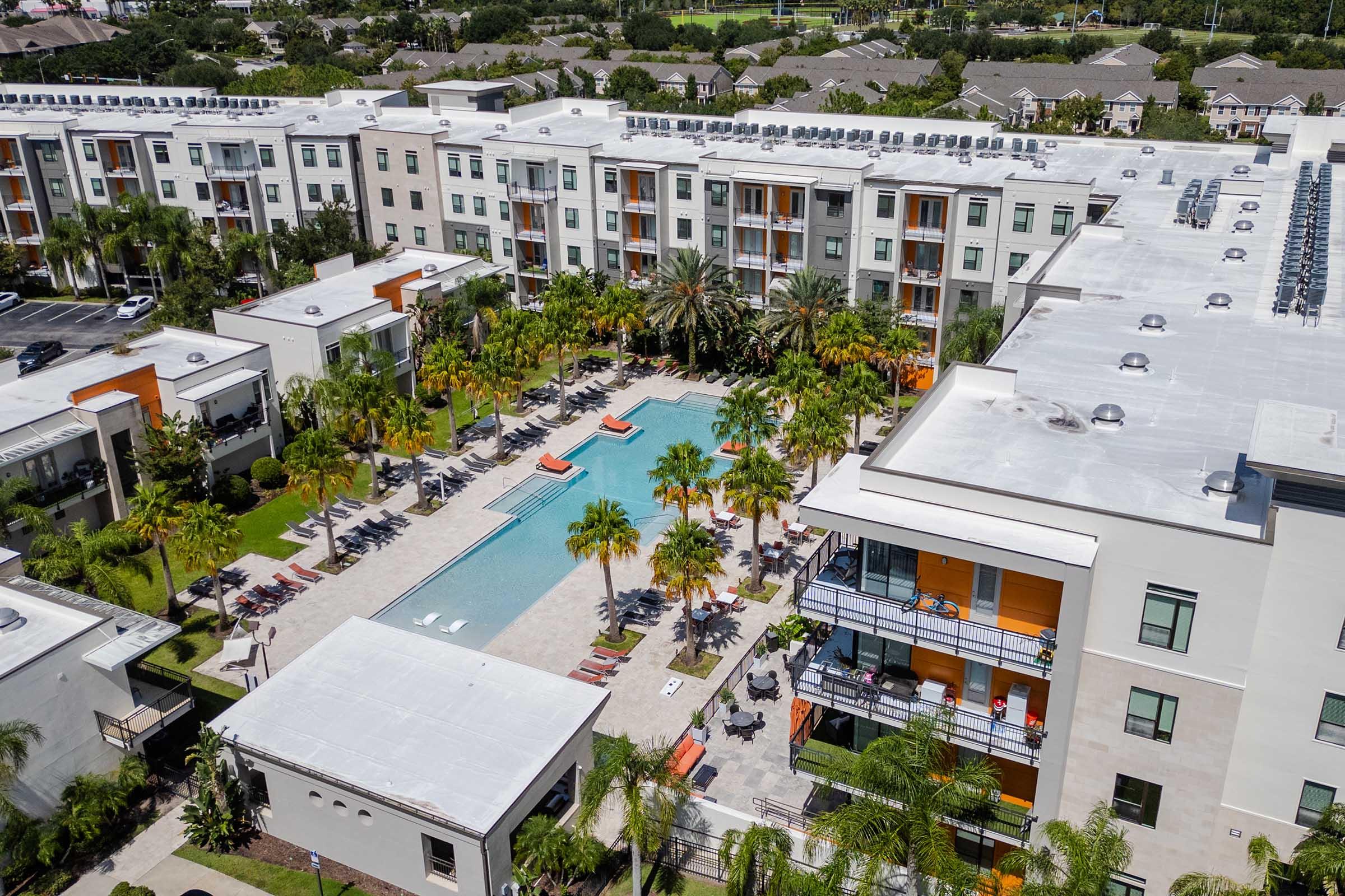 Aerial view of a modern apartment complex featuring a central swimming pool surrounded by palm trees, sun loungers, and patio areas. The buildings are contemporary in design with balconies and vibrant colors, set amidst landscaped gardens. Additional apartments are visible in the background.